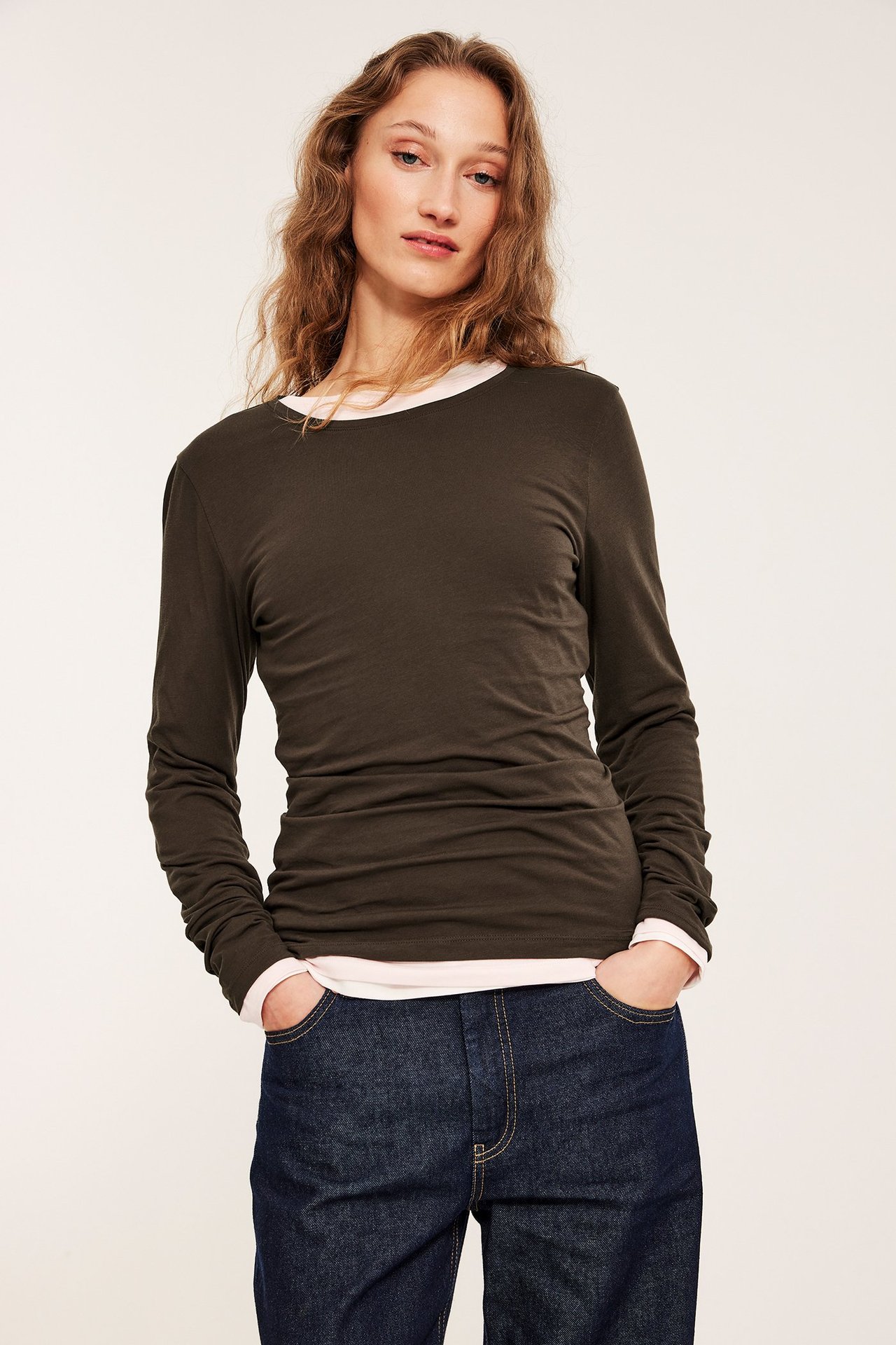 Frontbilde av en kvinne i en mørkebrun langermet basetopp med rund hals, over en lys rosa topp og mørkeblå jeans.