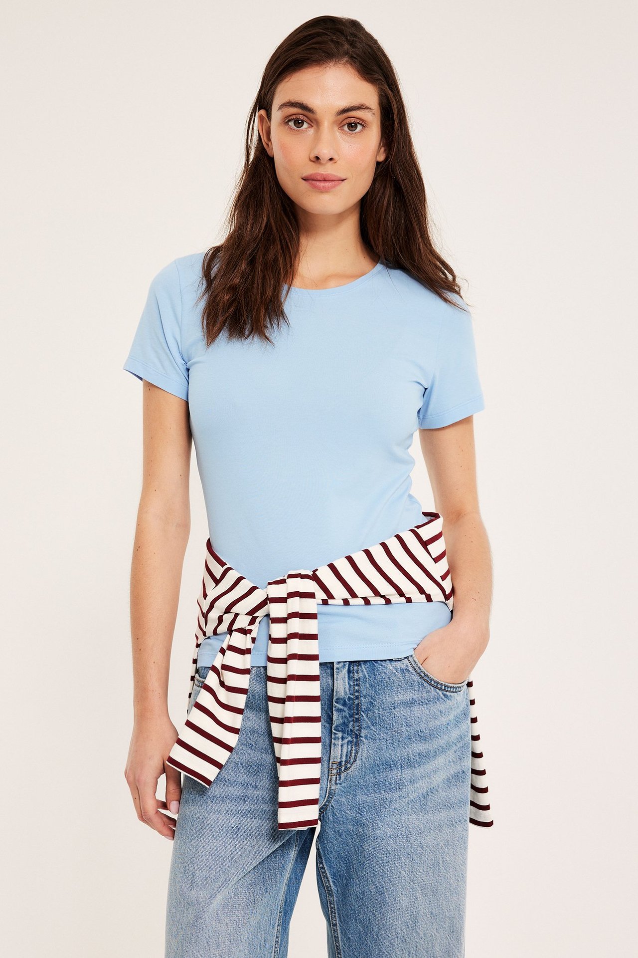 Frontvisning: Kvinne i lyseblå kortermet, rundhalset topp, jeans, og stripete genser knyttet i midjen.