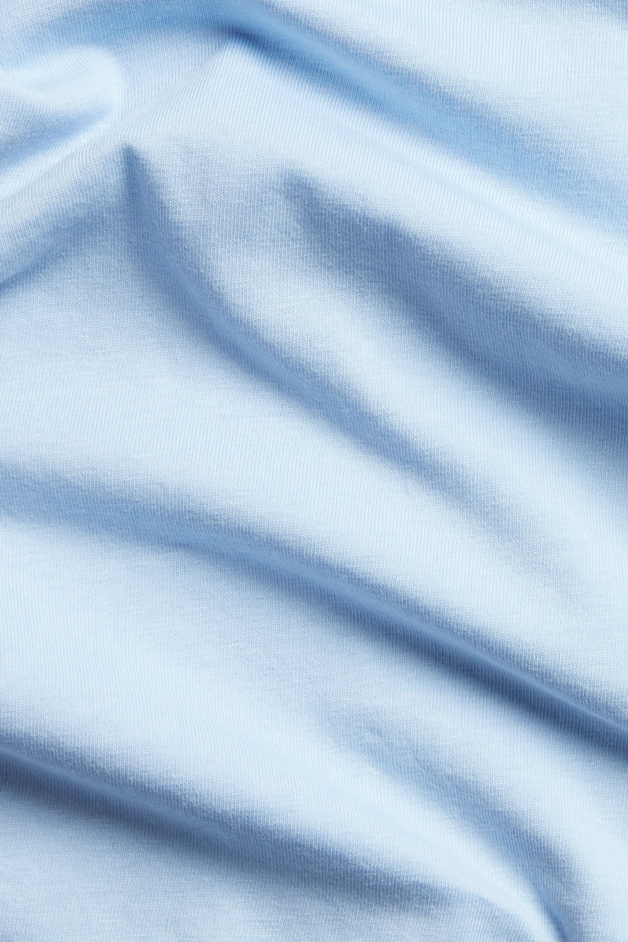 Zbliżenie jasnoniebieskiego, jednolitego materiału basic topu z bawełny, ukazujące teksturę i fałdy.