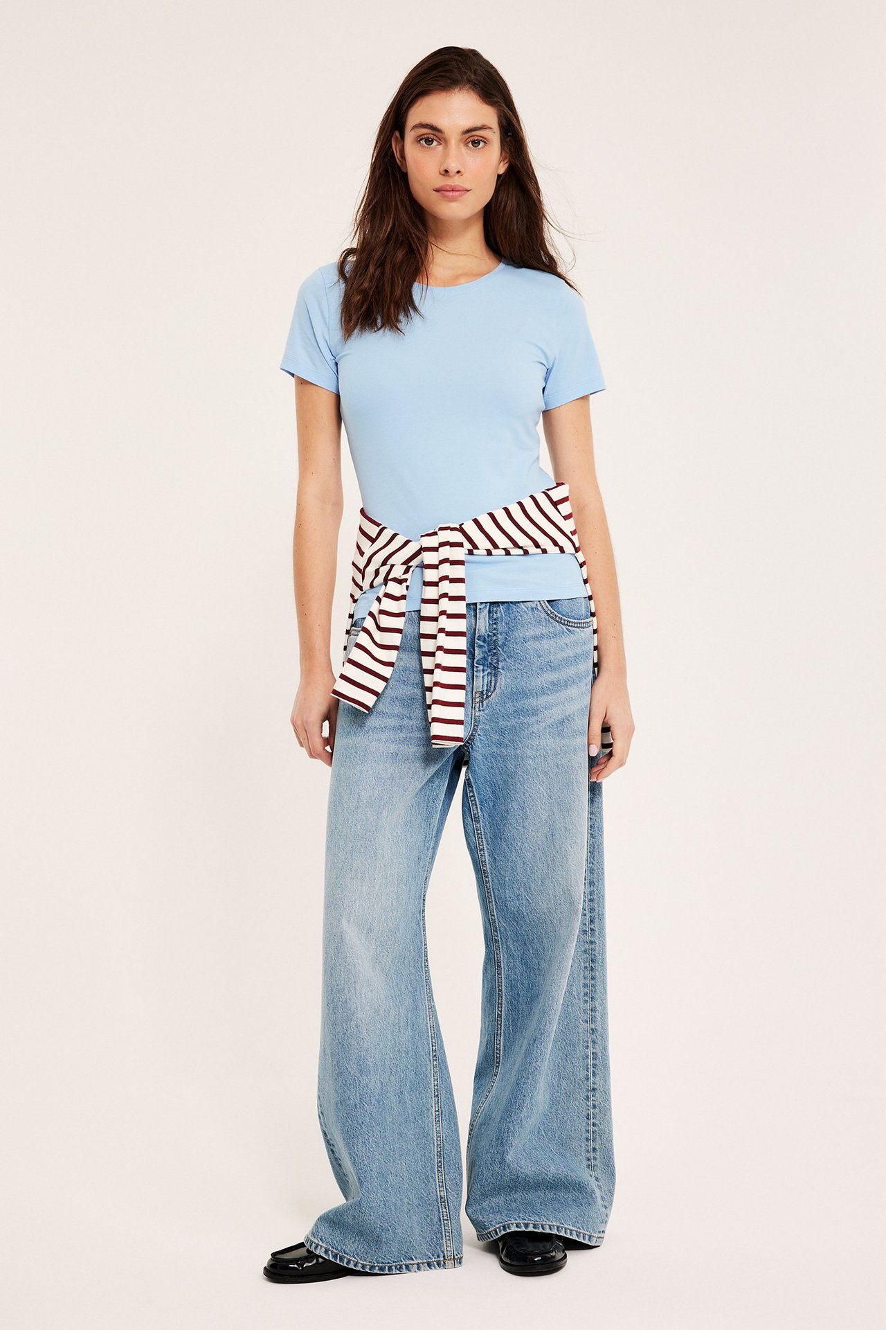 Forfra, kvinne i lyseblå kortermet basetopp, lyseblå vide jeans og en stripet genser knyttet rundt midjen.