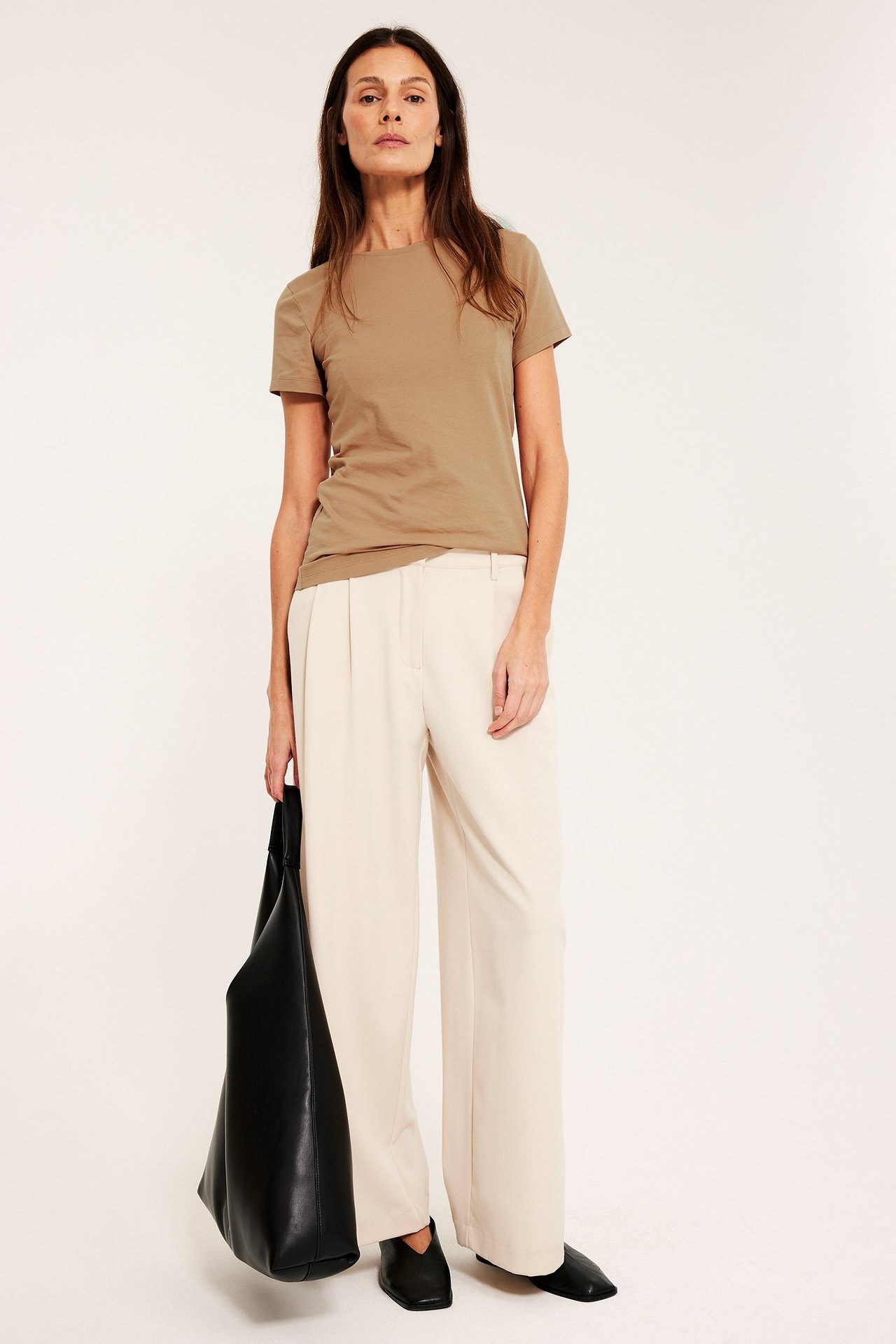 Edestä: Nainen beigessä lyhythihaisessa peruspaidassa, kermanvärisissä leveälahkeisissa housuissa, mustissa kengissä, pitäen mustaa laukkua.