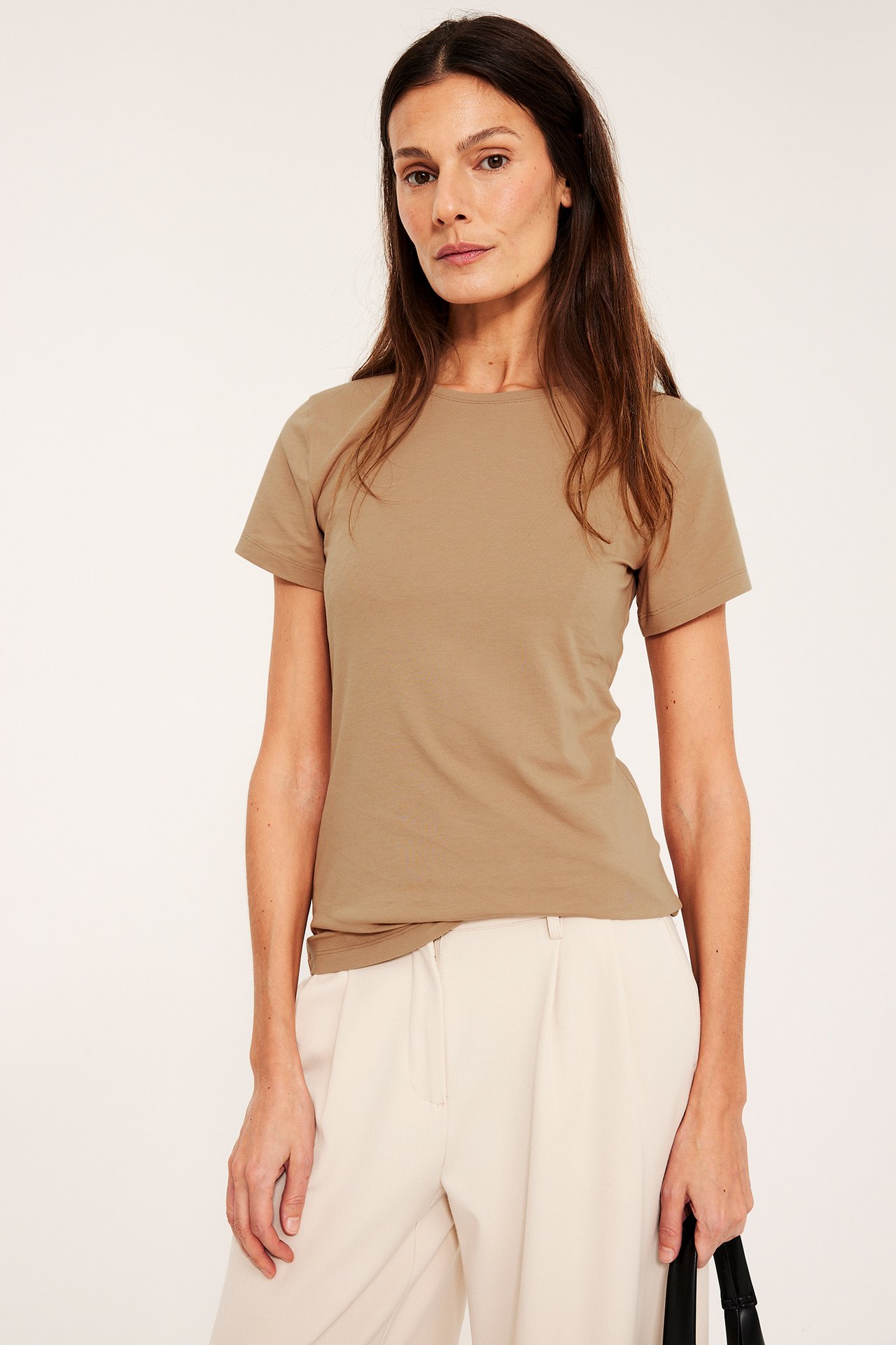 Edestä: nainen beigessä istuvassa lyhythihaisessa peruspaidassa, vaaleissa housuissa, pitelee mustaa käsilaukkua.