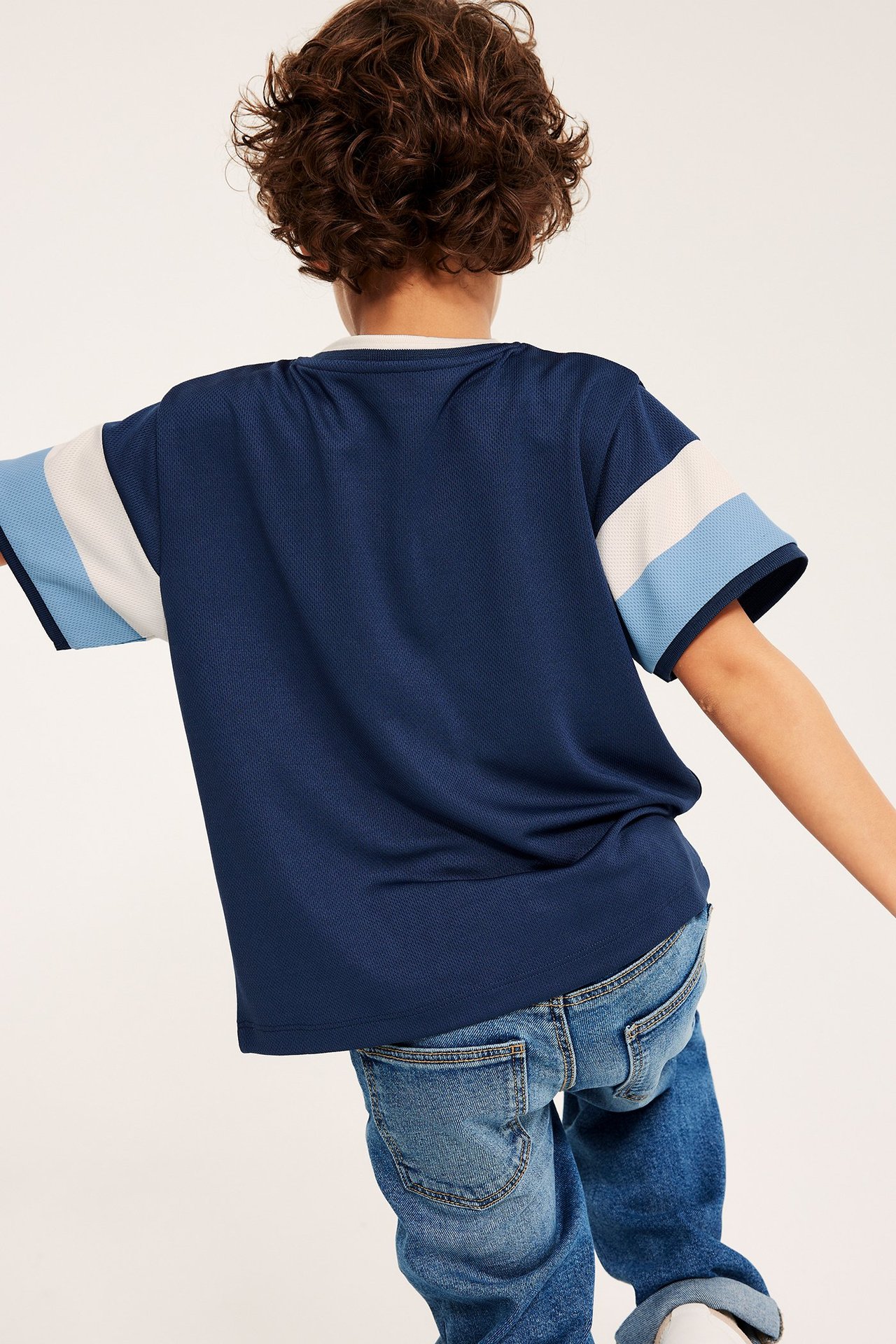 Bakfra, et barn i en marineblå mesh T-skjorte med lyseblå og hvite stripete ermer og blå jeans.