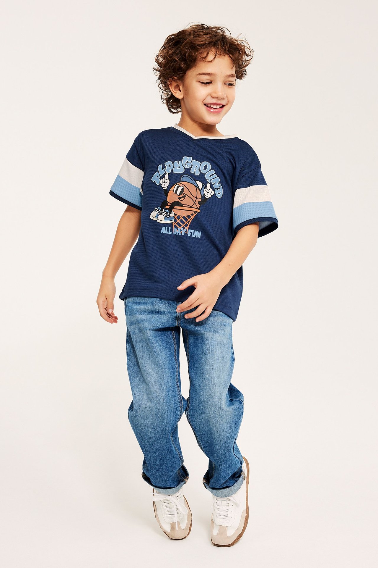 Edestäpäin, lapsi yllään laivastonsininen verkko-T-paita etupainatuksella, vaaleansiniset farkut ja valkoiset tennarit.