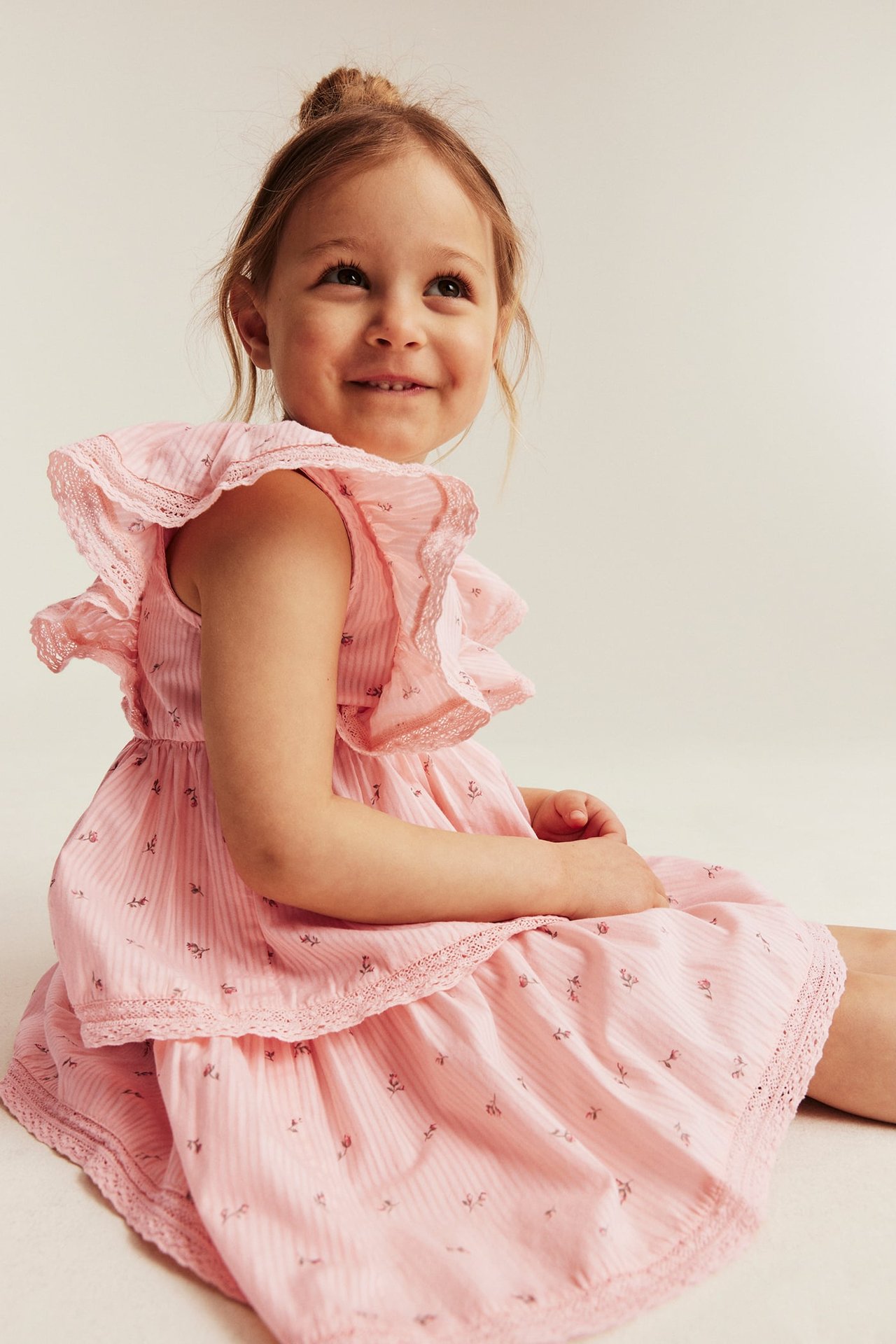 Widok z boku dziecka w jasnoróżowej sukience w kwiaty z paskami, falbankami i koronkowymi detalami.