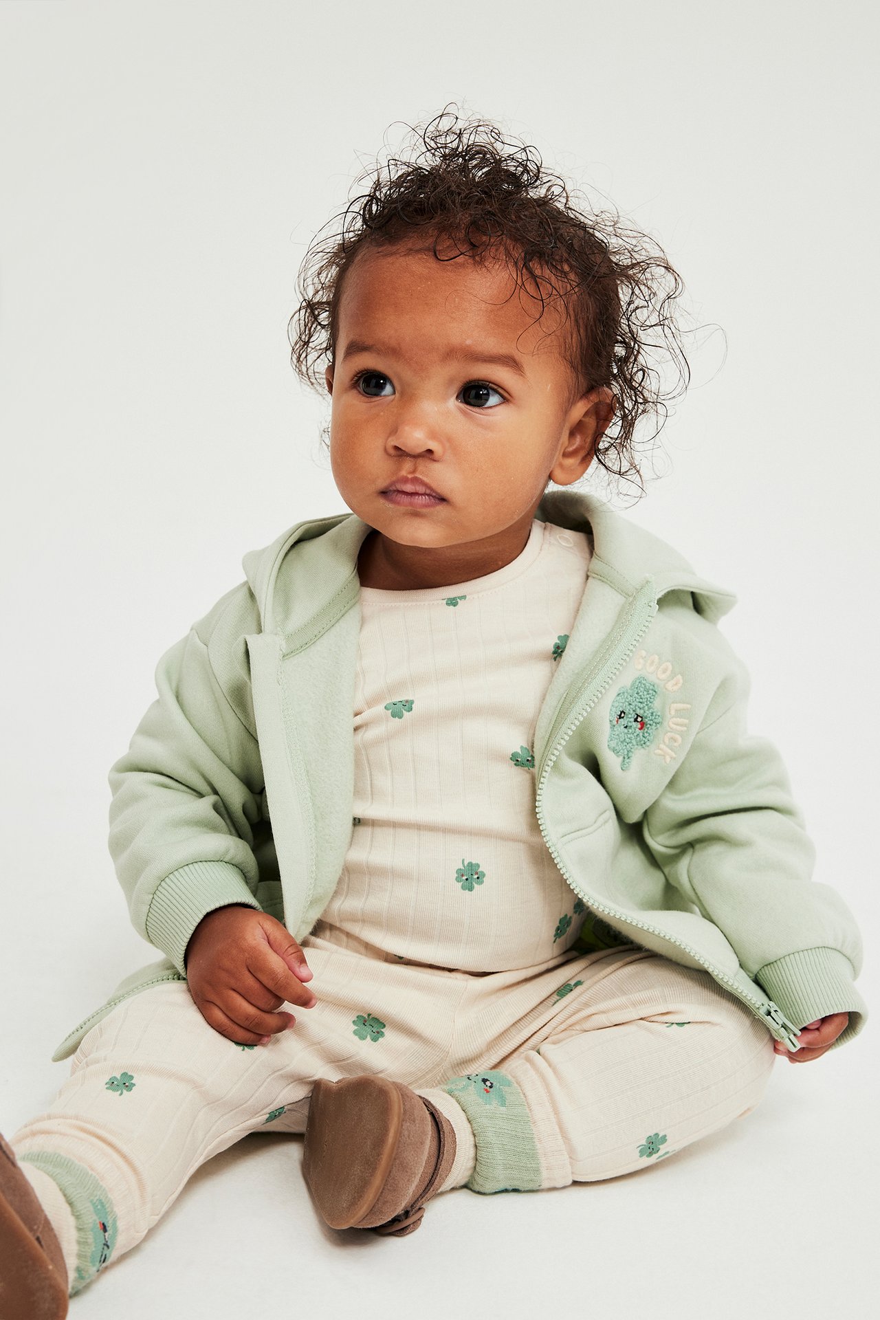 Helbild: Baby i beige ribbad långärmad body med klövermönster, under ljusgrön huvtröja och bruna skor.