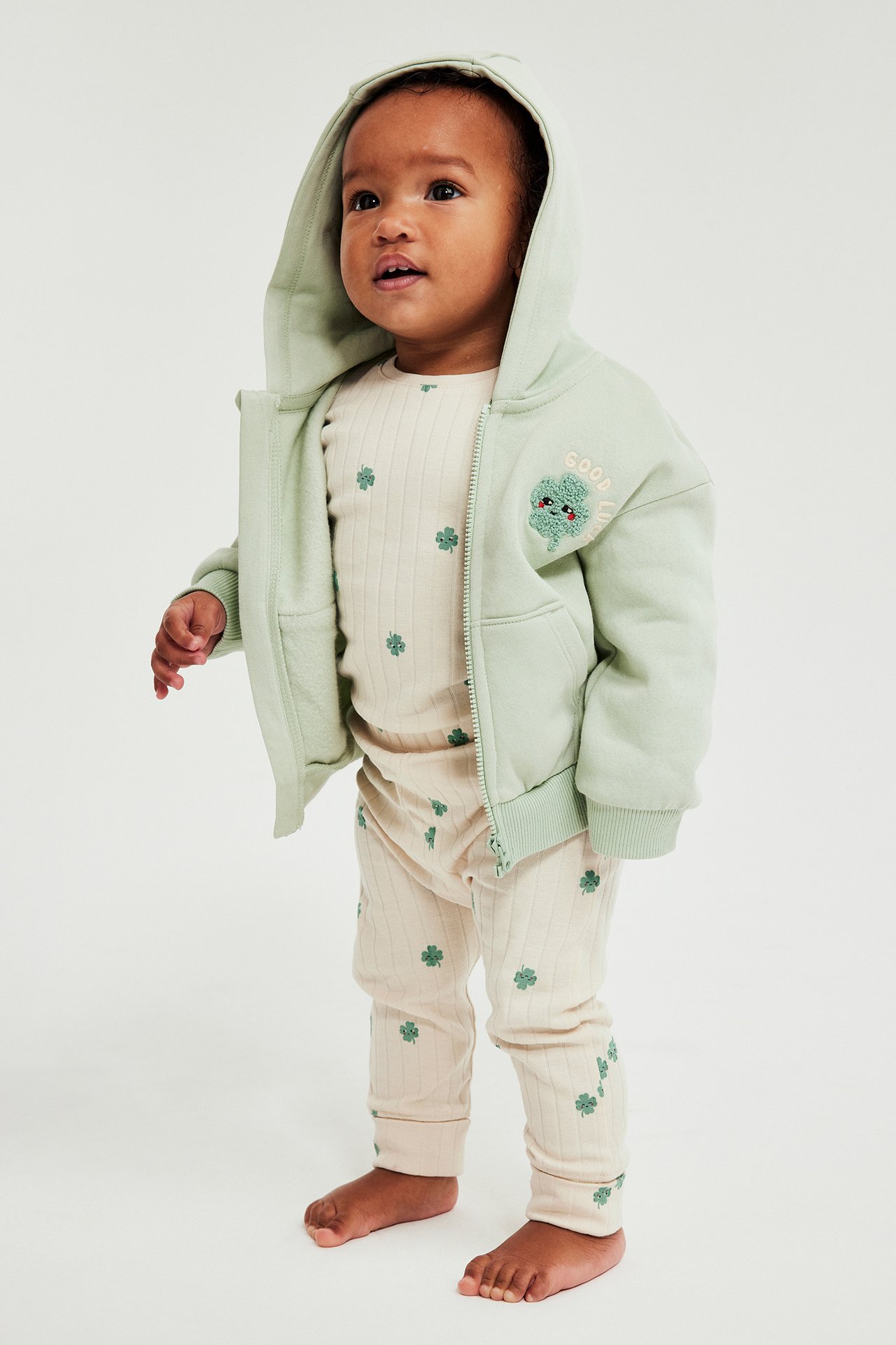 Framifrån: Baby i beige ribbad långärmad body med klövermönster, ljusgrön huvjacka och matchande leggings.