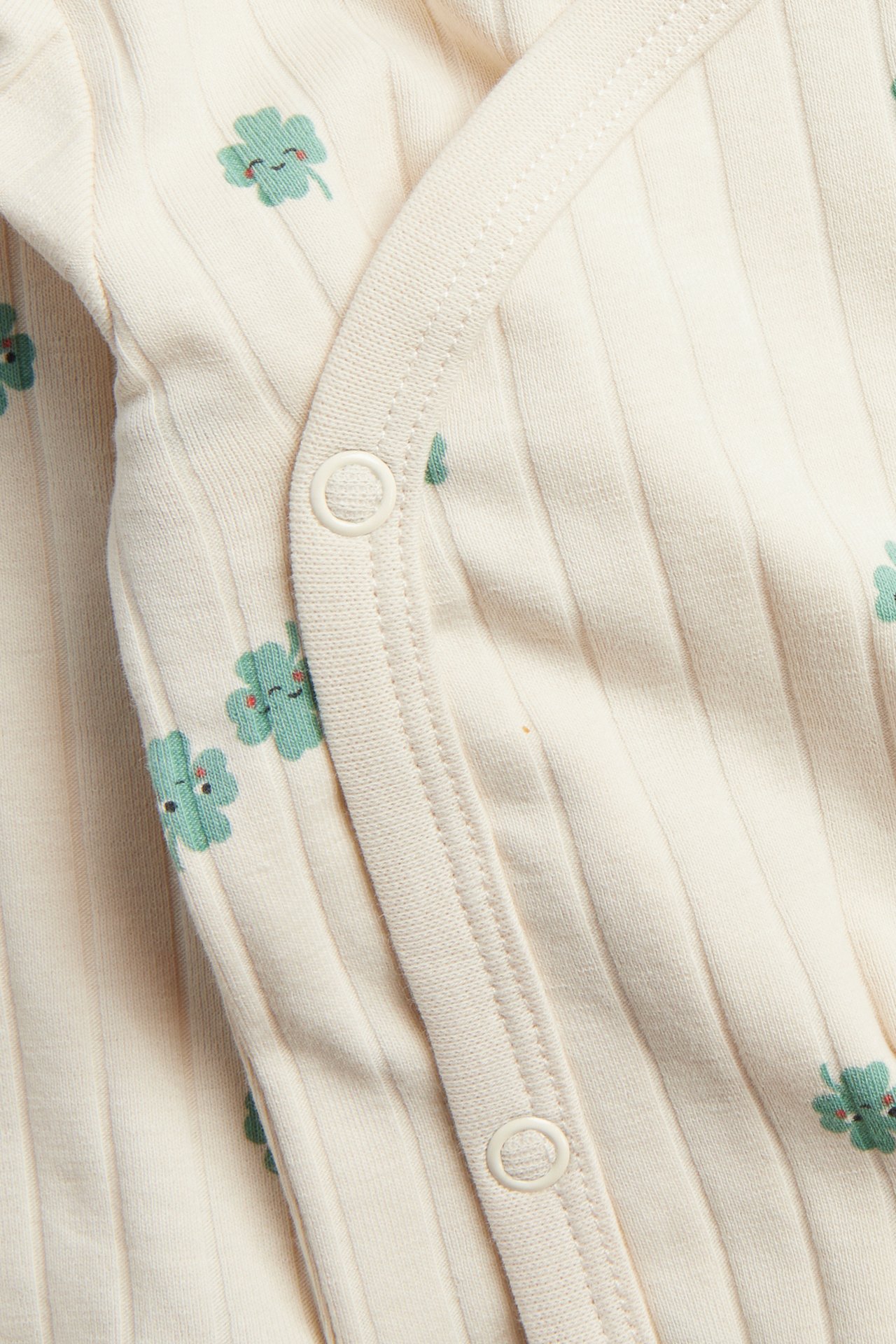 Närbild på en beige ribbad babybody med gröna klövertryck och vita tryckknappar.