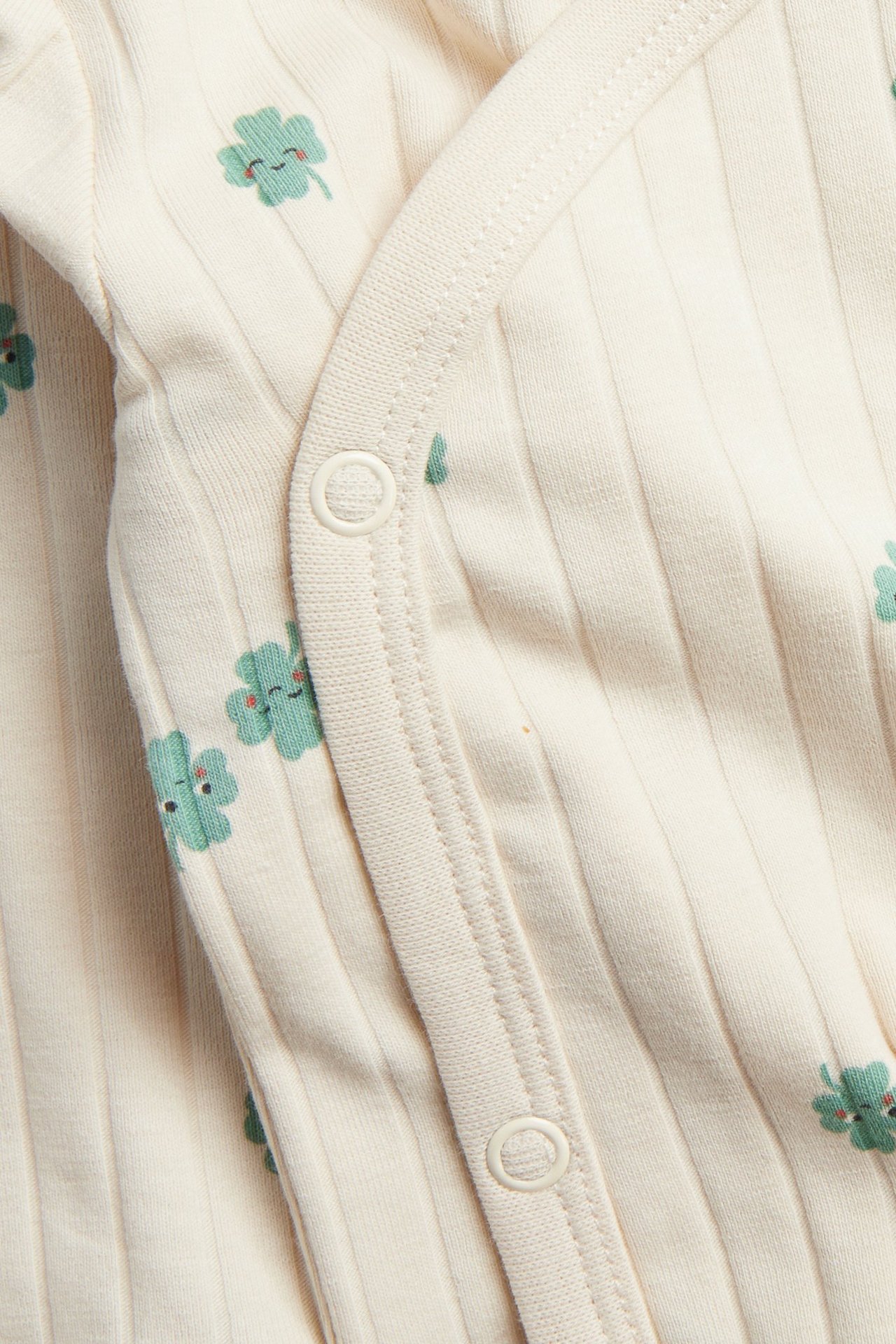 Zbliżenie na beżowe prążkowane body niemowlęce z zielonym nadrukiem koniczynek i białymi zatrzaskami.