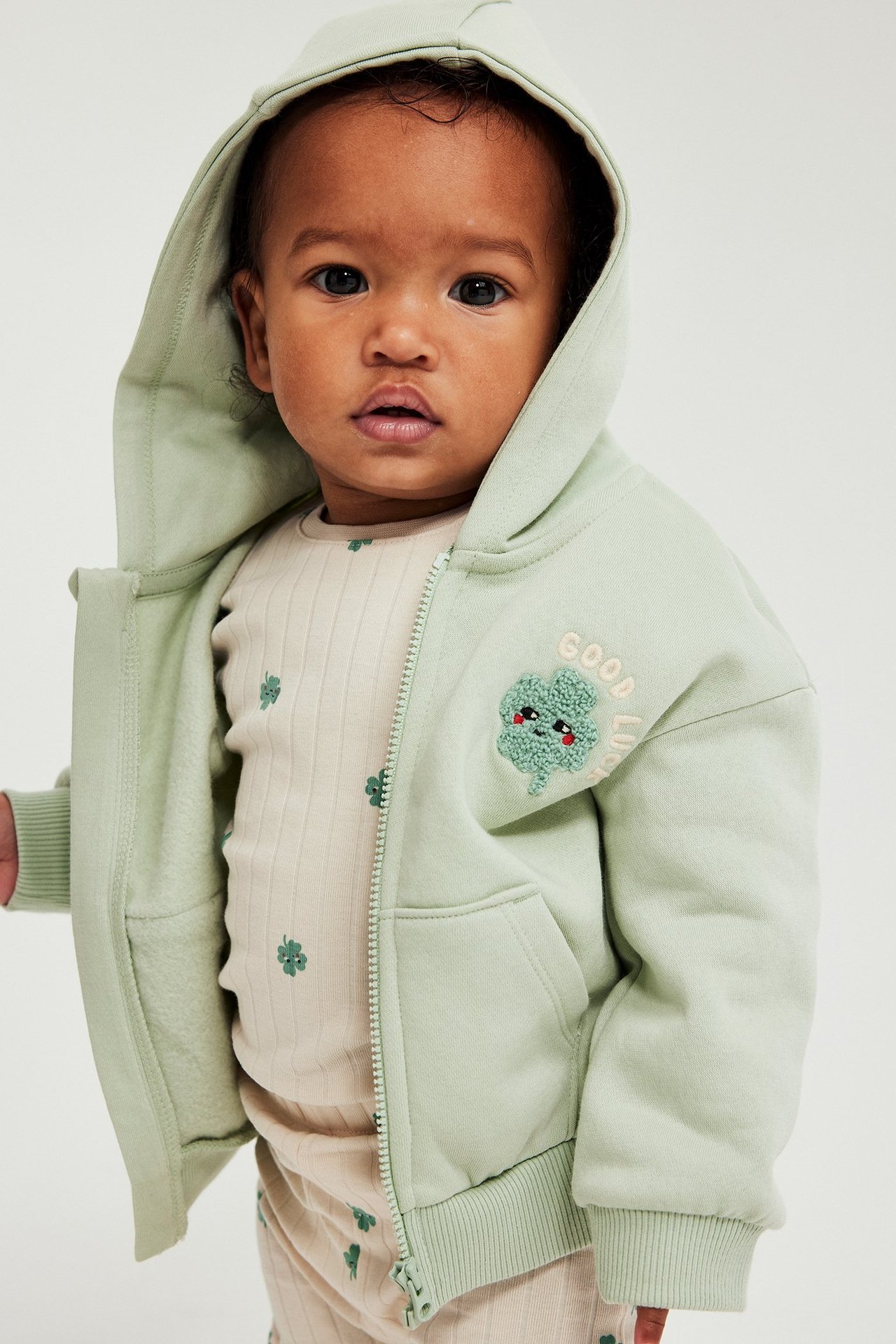 Närbild framifrån: baby i en beige ribbad body med klövermönster under en ljusgrön huvjacka med dragkedja.