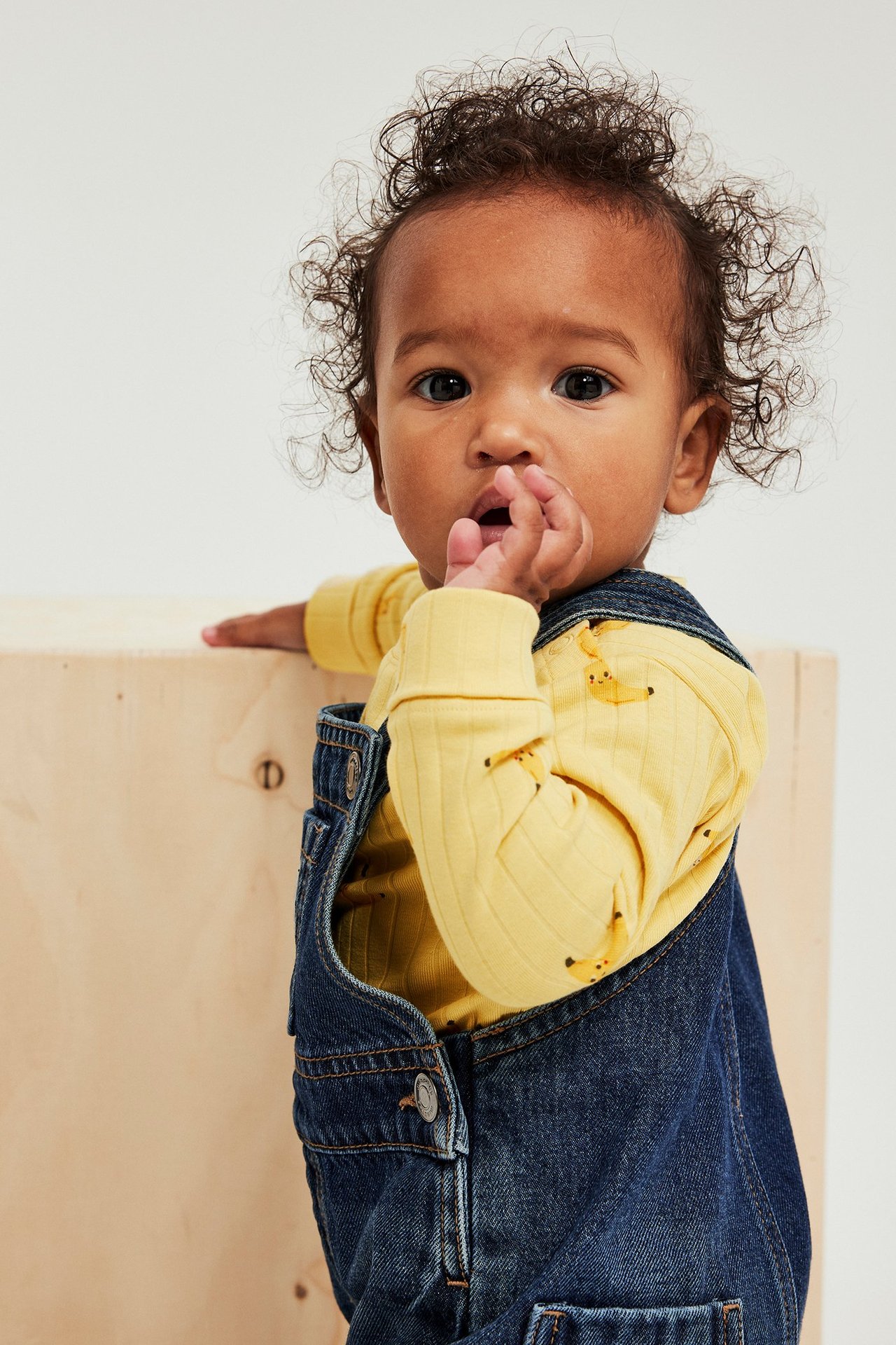 Lähikuva vauvasta keltaisessa ribatussa pitkähihaisessa bodyassa, jossa banaanikuvio, sinisten farkkuhaalareiden alla.