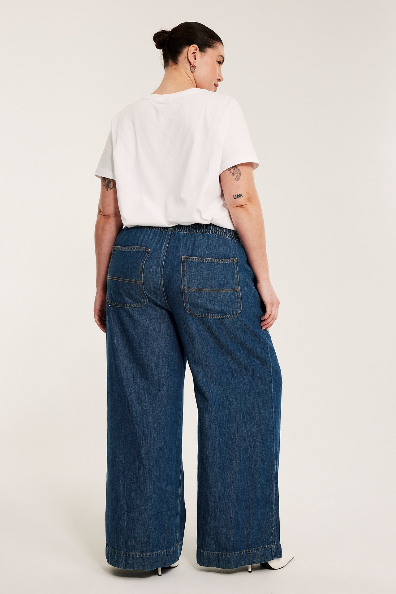 Naisen selkä, yllään tummansiniset leveät nyörilliset farkut takataskuilla ja valkoinen t-paita.