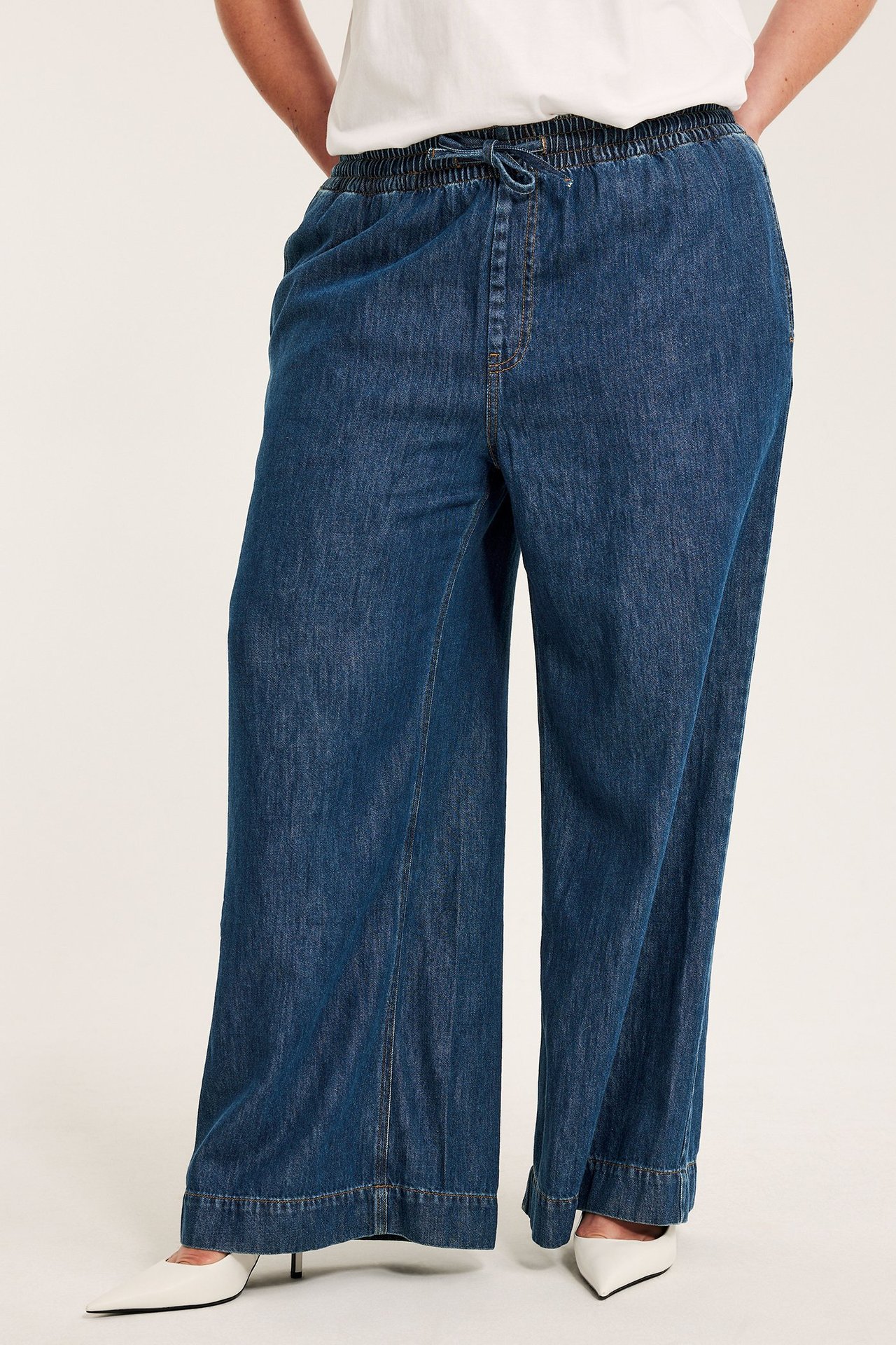 Edestä: Nainen tummansinisissä leveälahkeisissa pull-on-farkuissa kiristysnyörillä, valkoisessa t-paidassa ja valkoisissa kärkikoroissa.