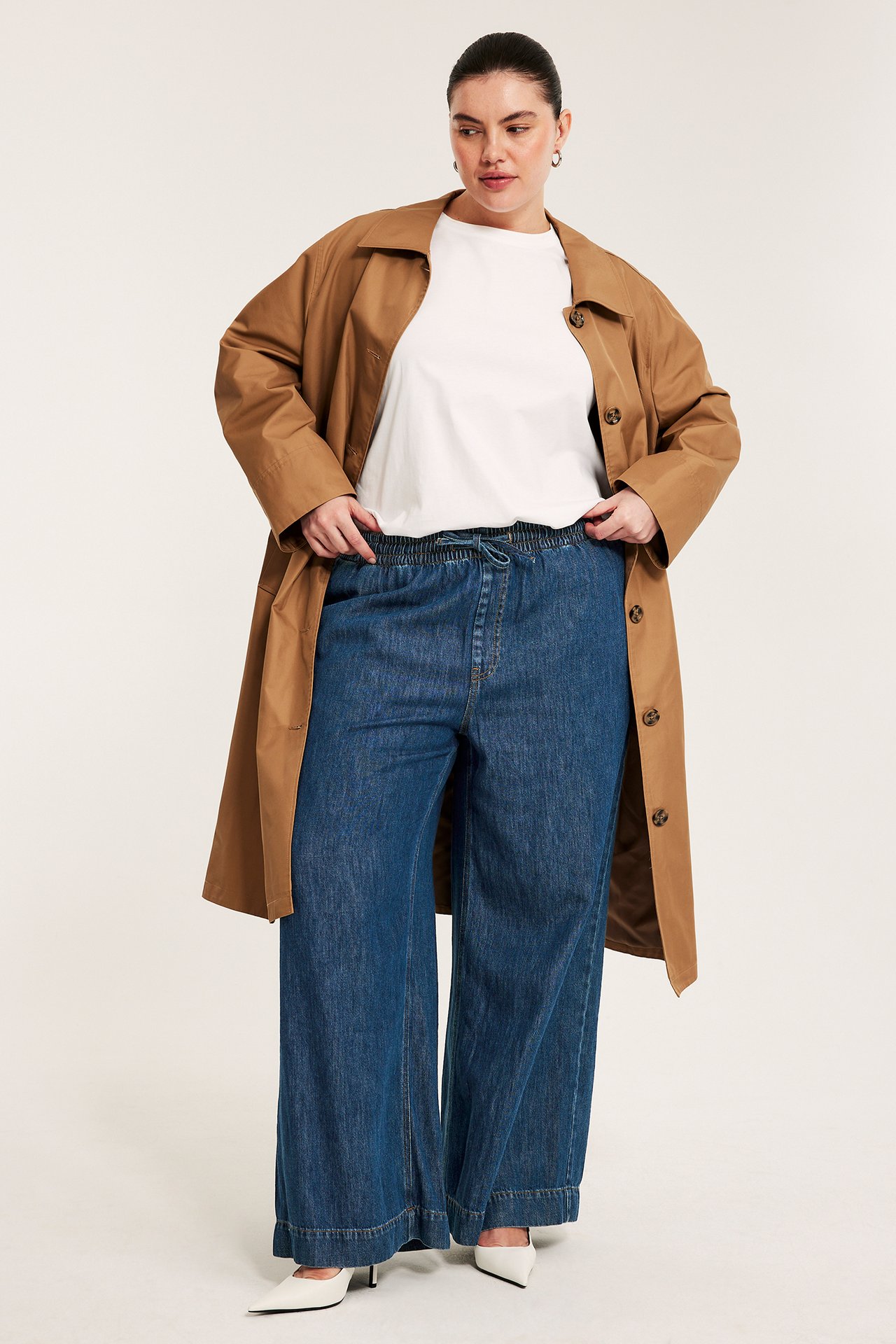 Naisen etunäkymä tummansinisissä leveissä farkuissa, kiristysnyörillä, valkoisessa t-paidassa ja ruskeassa trenssitakissa.