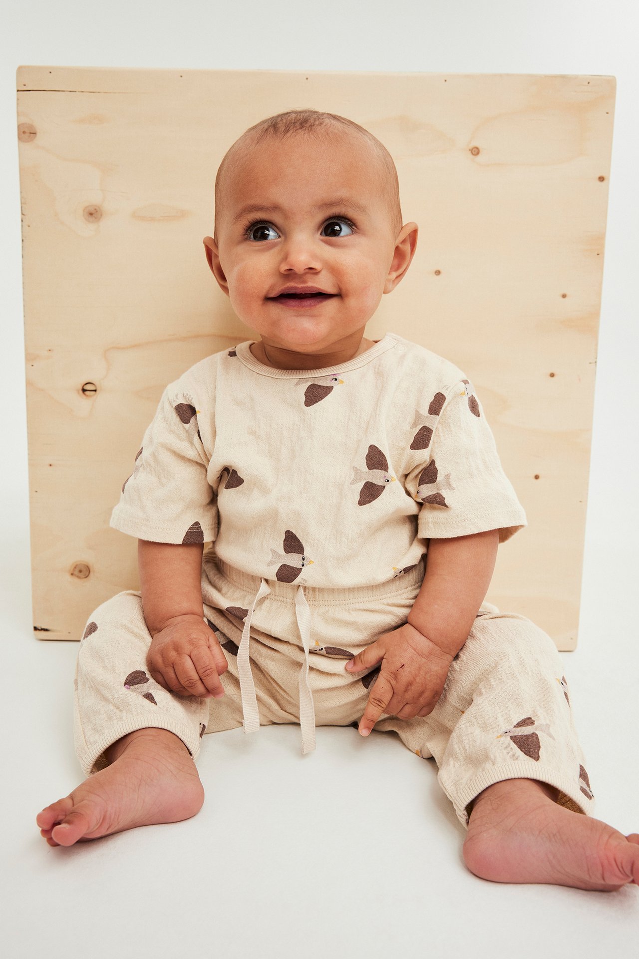 Framifrån, baby i beige mönstrad oversized body med tryckknappar och matchande byxor.