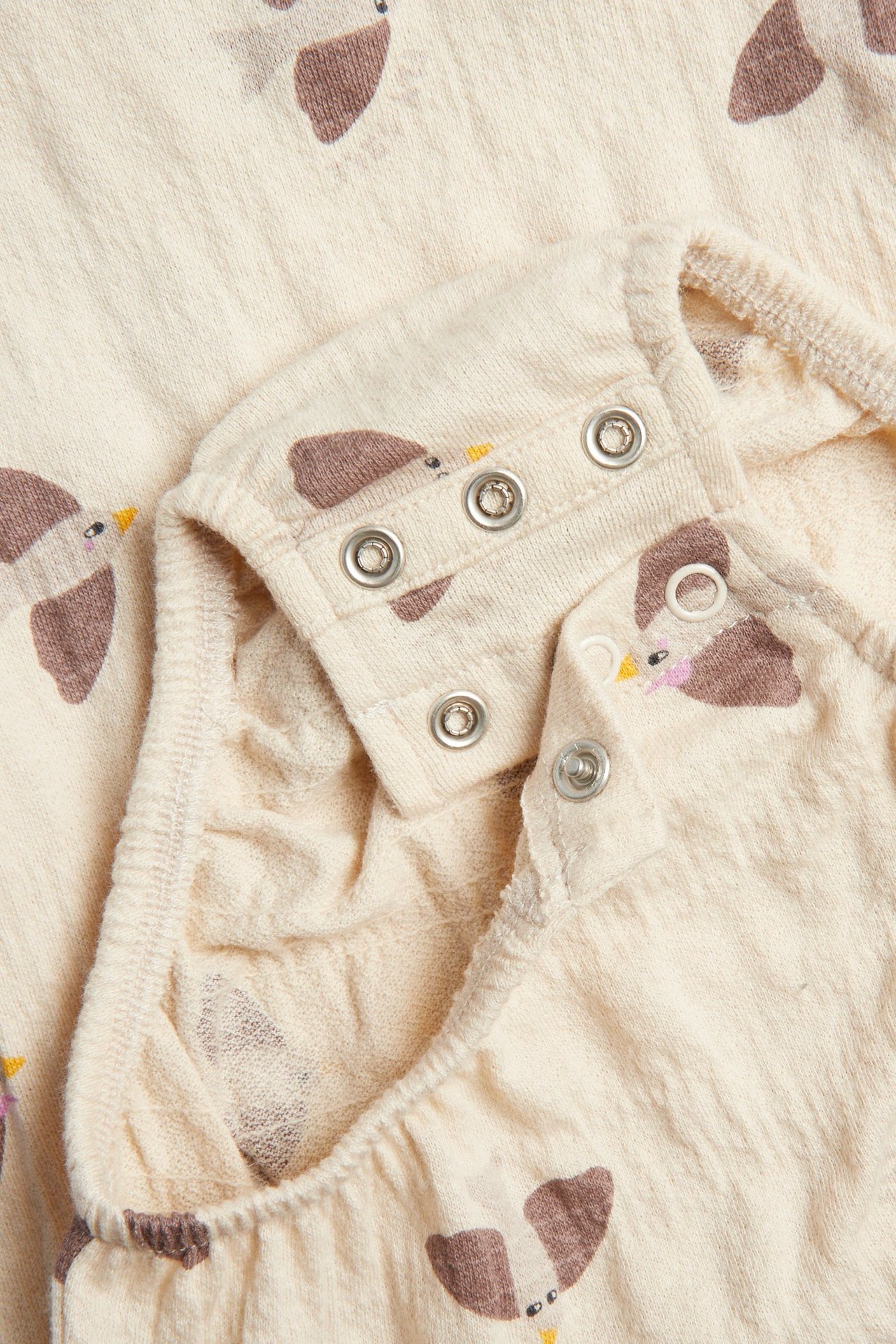 Närbild av en beige oversized babybody med brunt fågelprint och tryckknappar vid axeln.