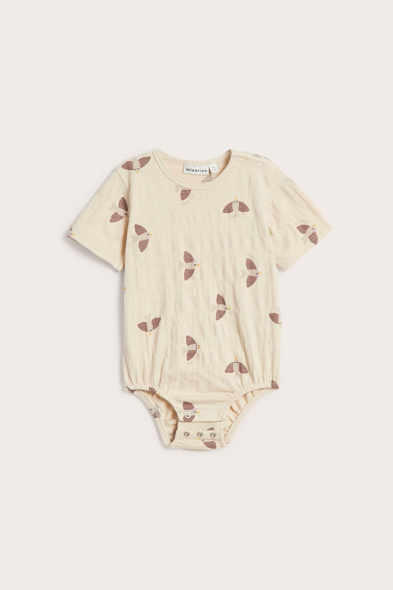 Frontvisning av en oversized beige babybody i bomull med fuglemønster og trykknapper.