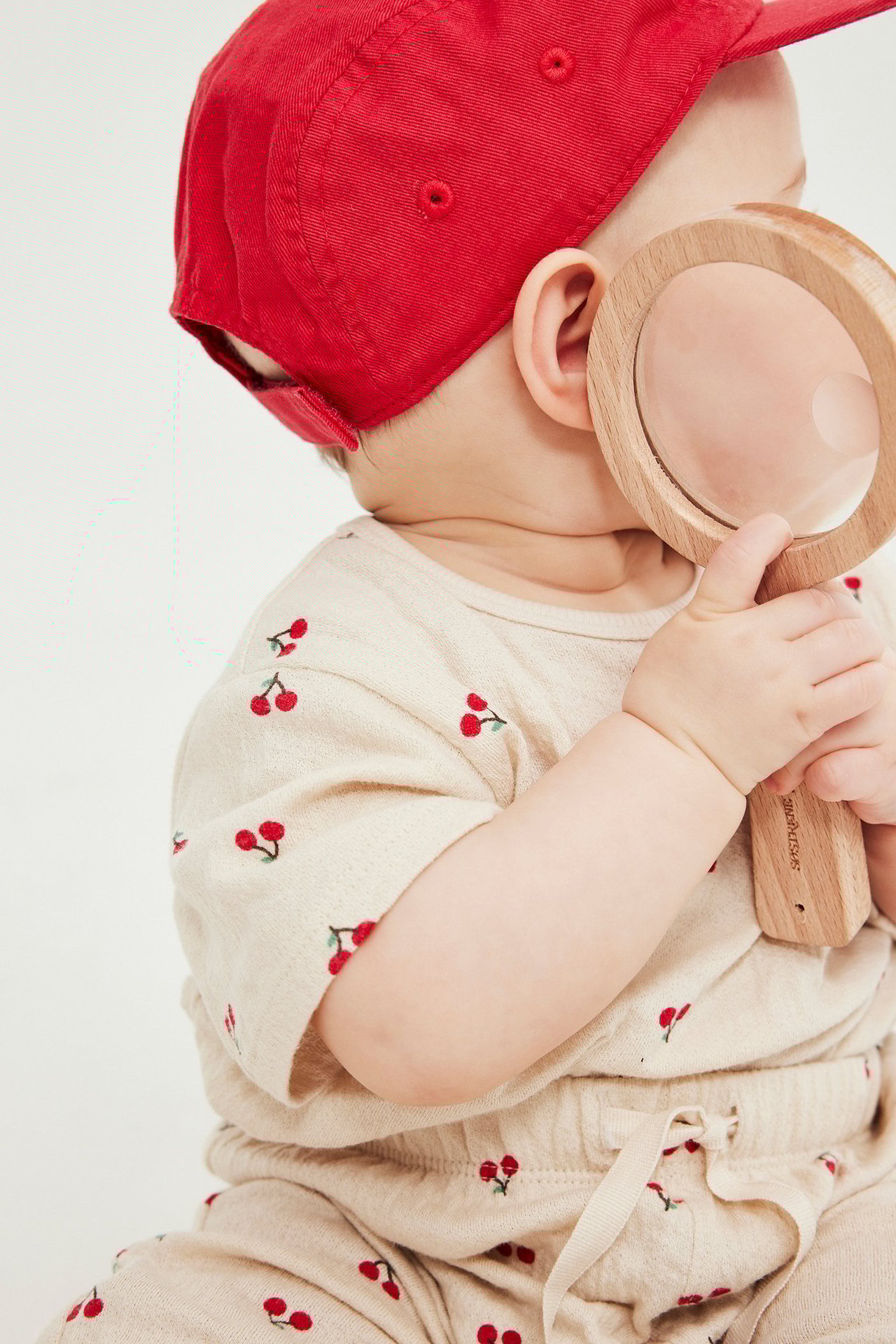 Zbliżenie z boku: Dziecko w jasnobeżowym body w wisienki, pasujące spodnie, czerwona czapka, trzyma lupę.