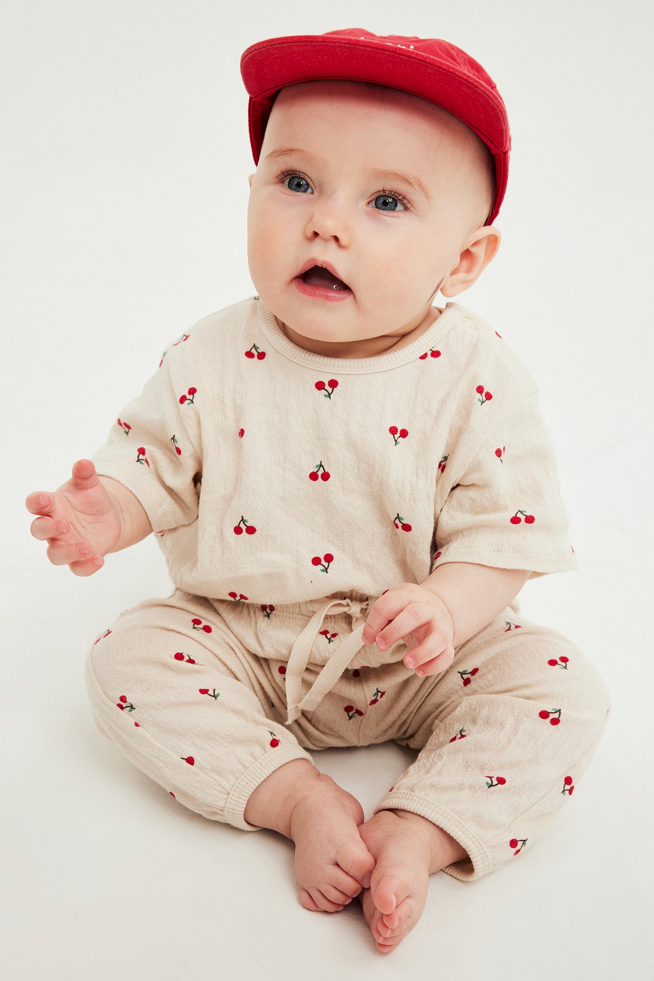 Från fronten, en baby som sitter i en ljusbeige oversized body med röda körsbär och en röd keps.