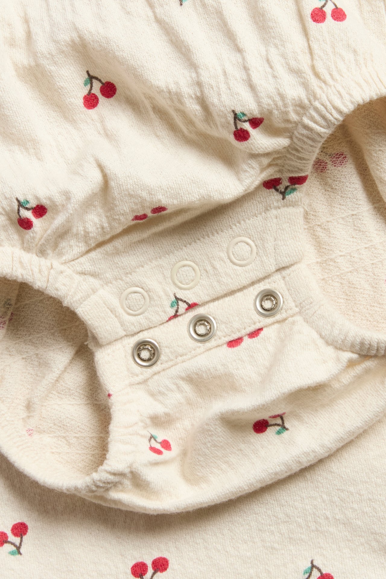 Zbliżenie beżowego body niemowlęcego oversize w czerwone wisienki z zatrzaskami.