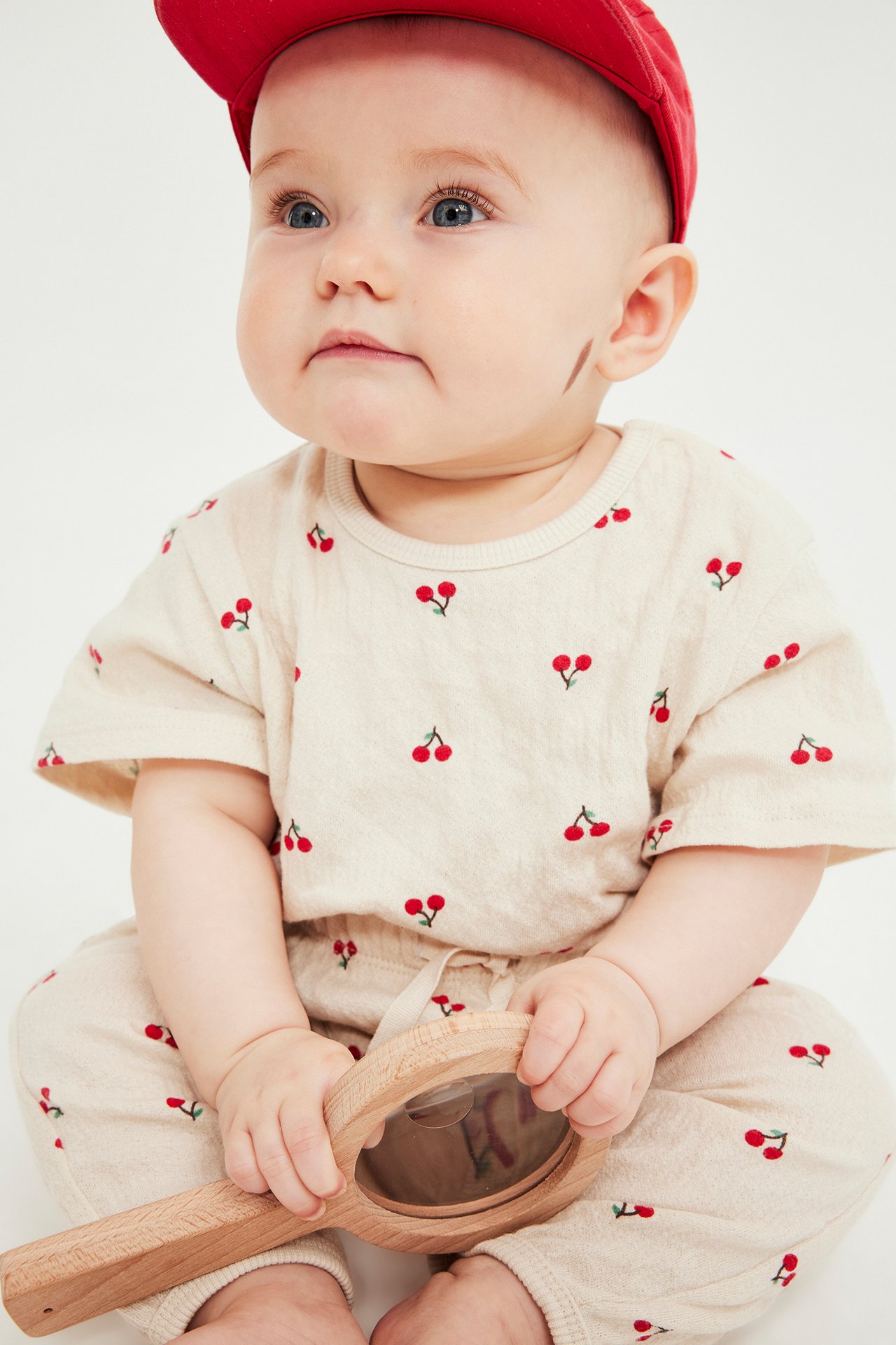 Edestä: vauva vaaleabeigessä kirsikkabodyssä, housuissa, punaisessa lippalakissa, puulelu kädessä.