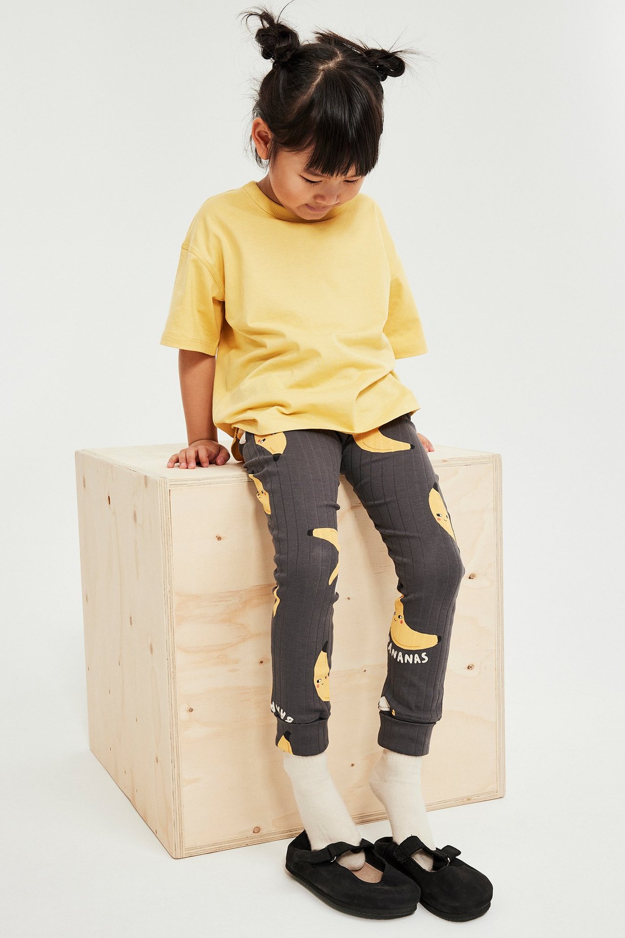 Framsida: Barn bär en lös gul t-shirt, gråa leggings med banantryck, vita strumpor och svarta skor.