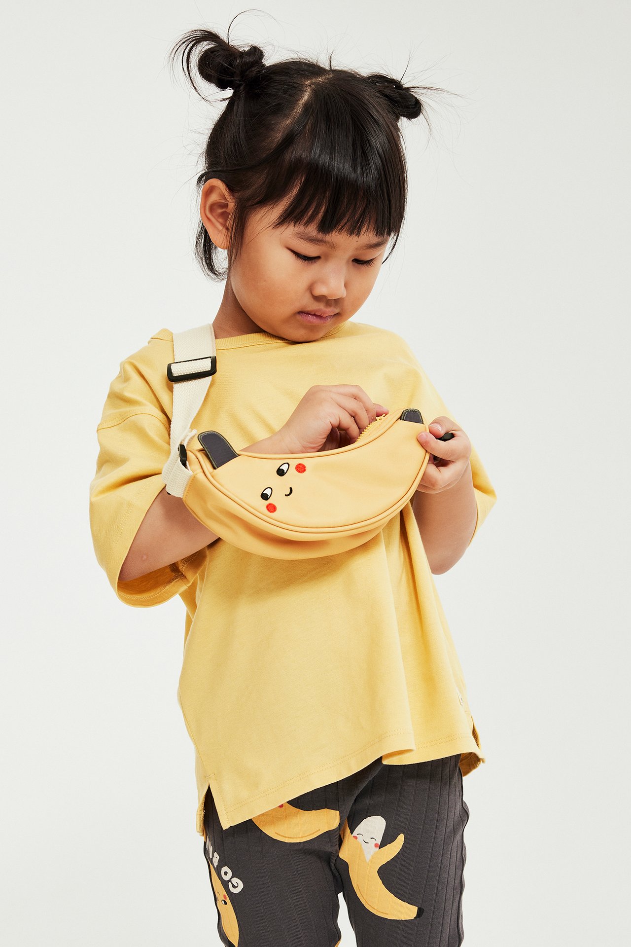 Framifrån, ett barn i gul bomulls-t-shirt, mörkgrå bananmönstrade leggings och med en gul banan-crossbodyväska.