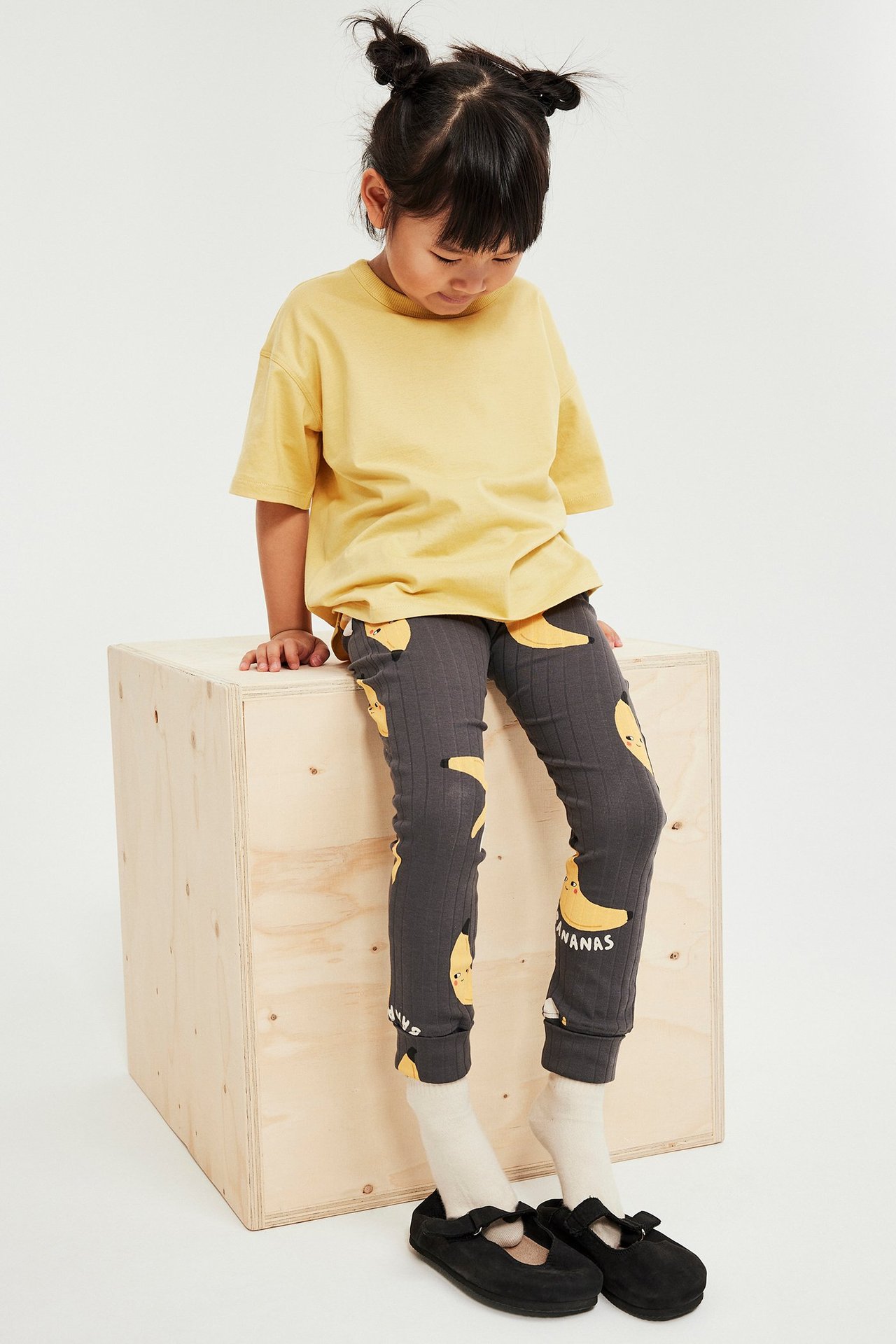 Framifrån: Ett barn i en lös gul bomulls-t-shirt, mörkgrå ribbade leggings med bananmönster och svarta skor.