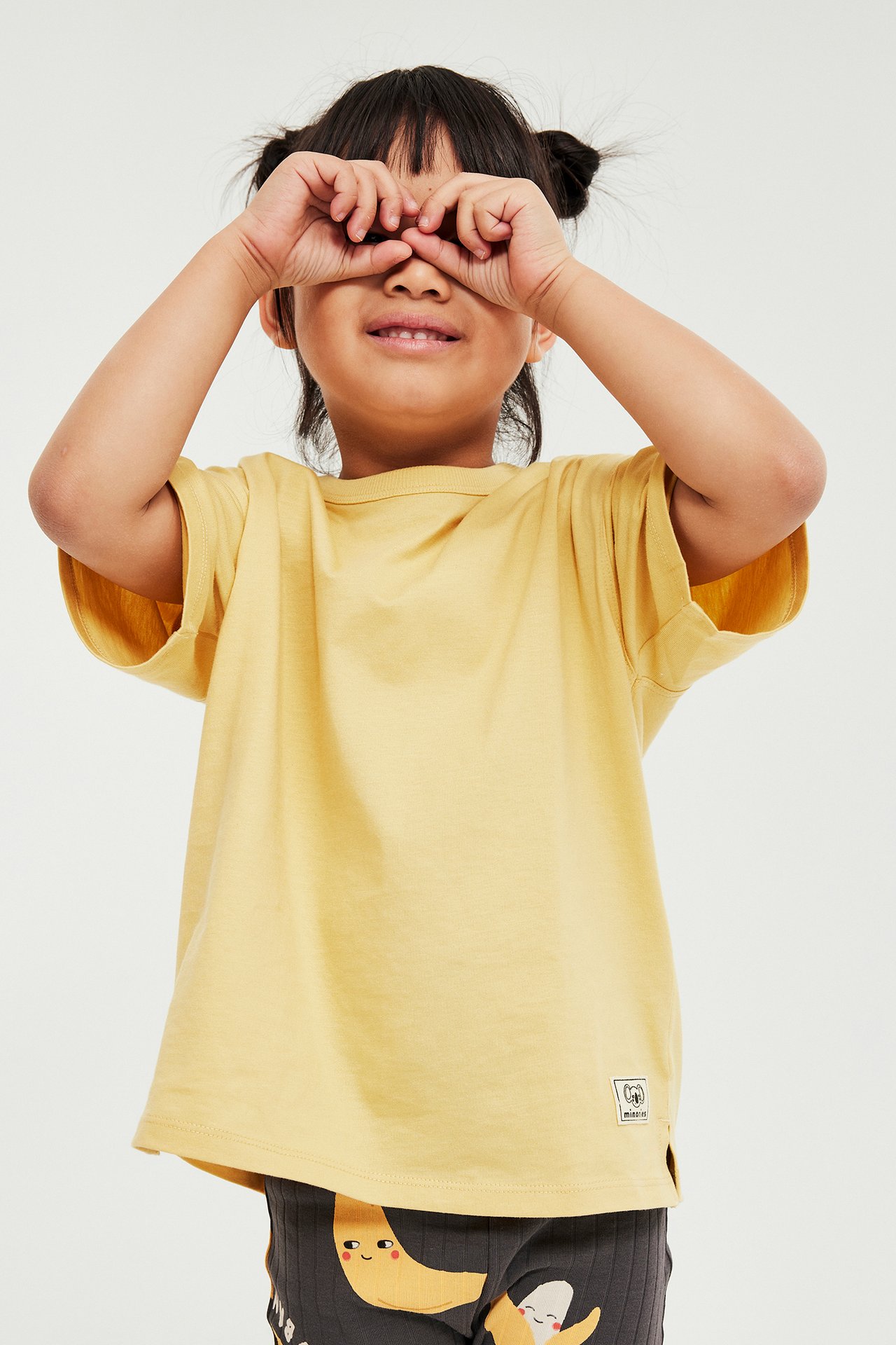 Framifrån: Barn i gul kortärmad bomulls-t-shirt med märke, mörka mönstrade leggings, händerna för ögonen.