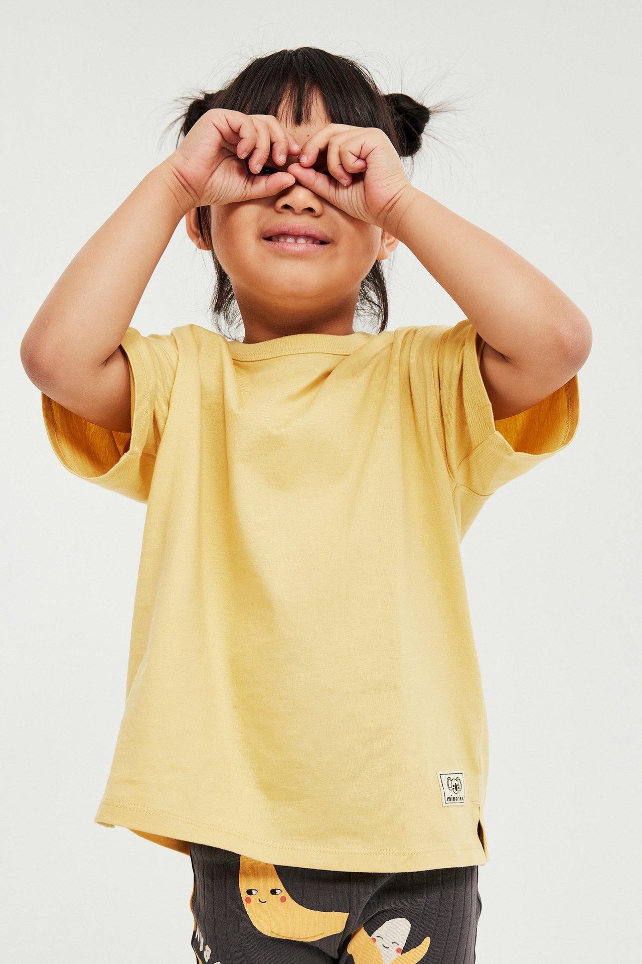 Widok z przodu: Dziecko w żółtej bawełnianej koszulce z krótkim rękawem z naszywką, ciemne legginsy, zakrywa oczy.