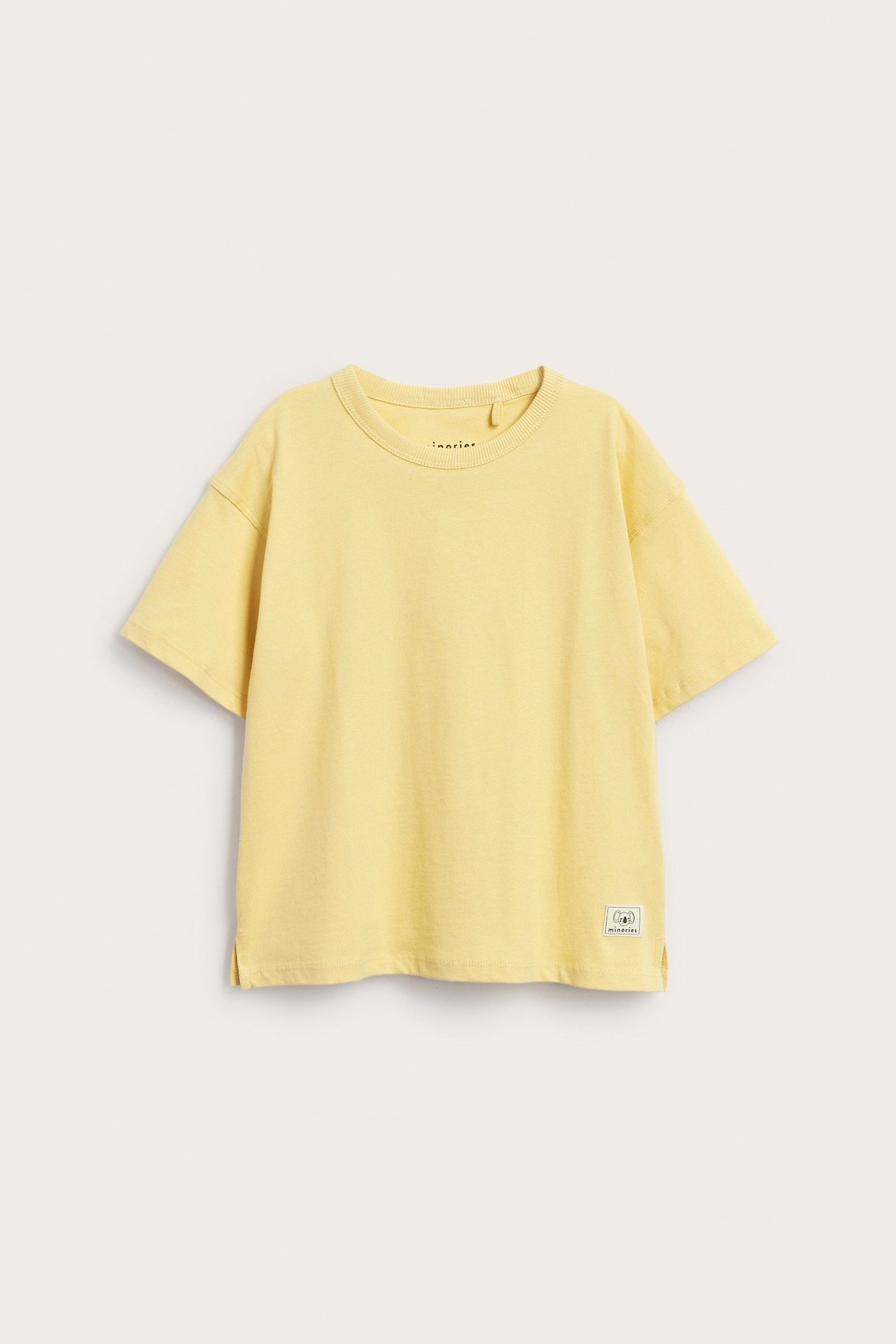 Framifrån: en lös, gul bomulls t-shirt för barn med korta ärmar och vävt märke.