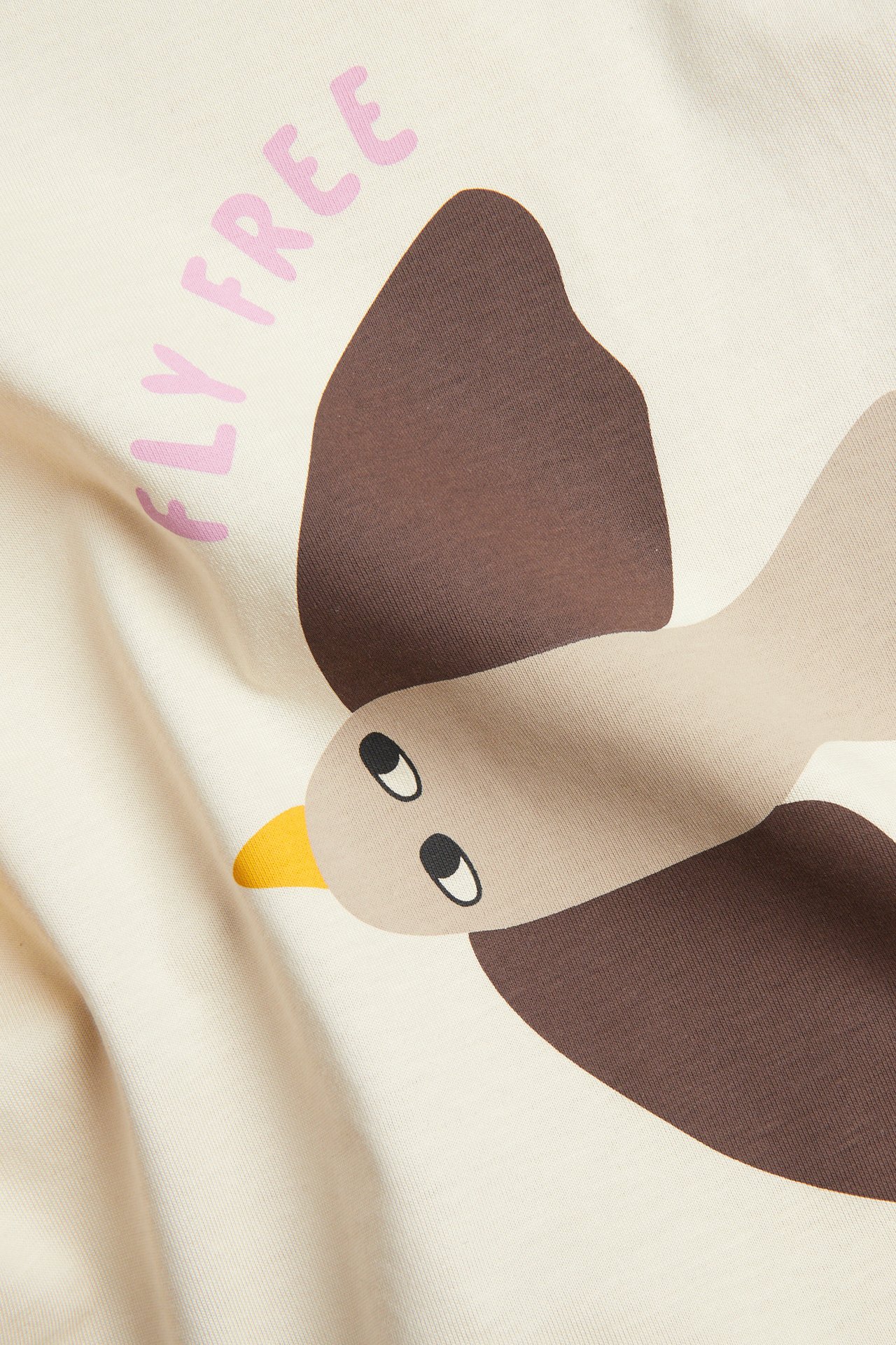 Lähikuva beigestä puuvilla-T-paidasta ruskealla lintupainatuksella ja vaaleanpunaisella 'FLY FREE' -tekstillä.