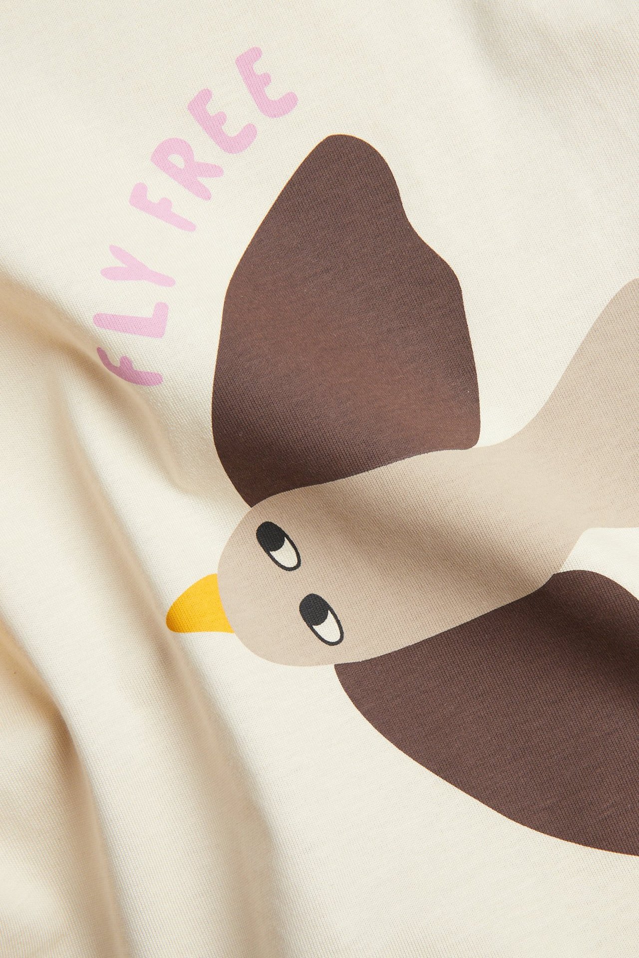 Zbliżenie na beżowy bawełniany T-shirt z krótkim rękawem, z brązowym nadrukiem ptaka i różowym napisem 'FLY FREE'.