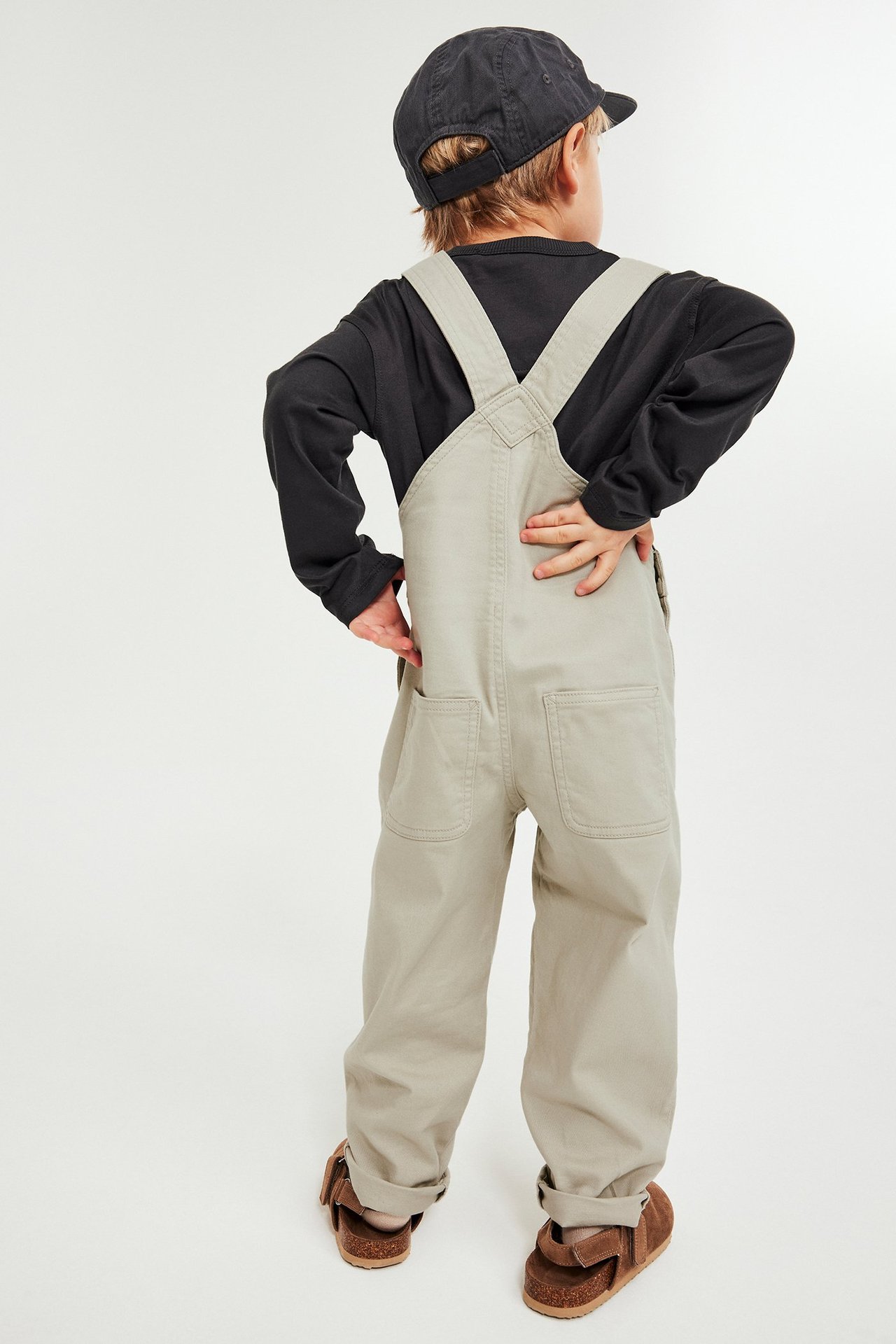 Lapsi takaapäin yllään beiget twill-haalarihousut mustan pitkähihaisen paidan, mustan lippiksen ja ruskeiden sandaalien kanssa.