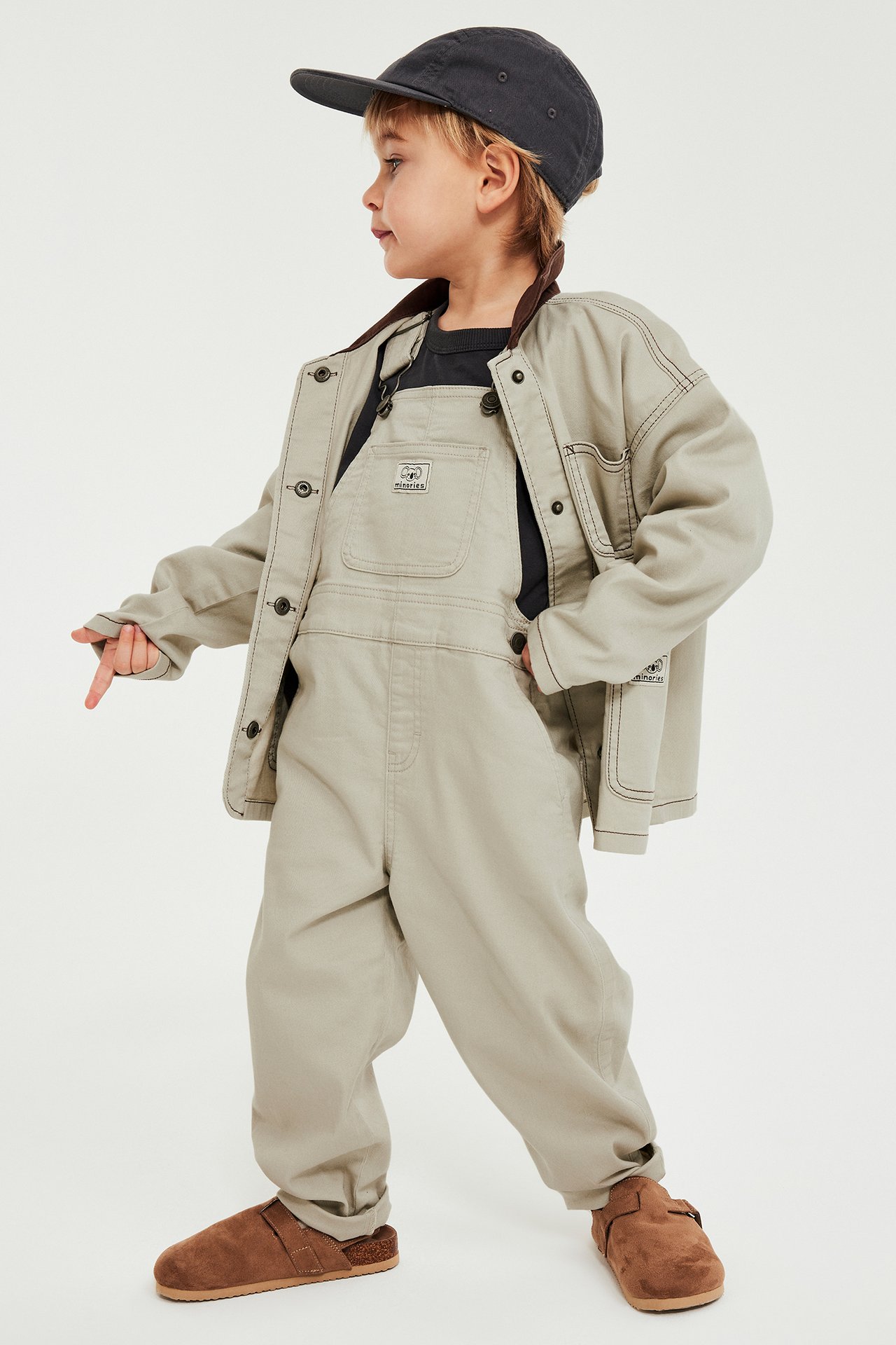 Lapsi sivukuva: beiget twill-haalareissa, musta t-paita, beige takki, harmaa lippis ja ruskeat tohvelit.