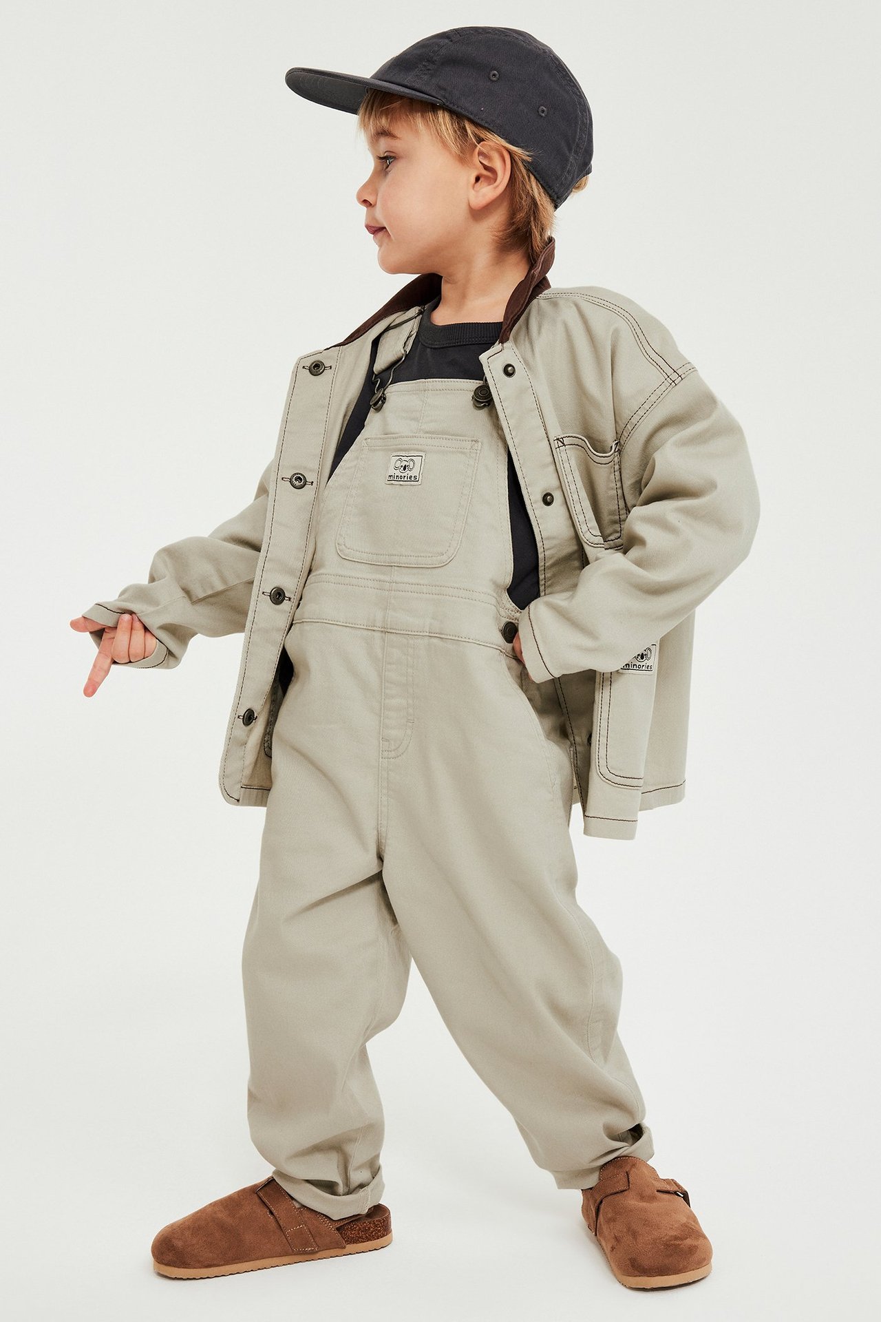 Lapsi sivukuva: beiget twill-haalareissa, musta t-paita, beige takki, harmaa lippis ja ruskeat tohvelit.