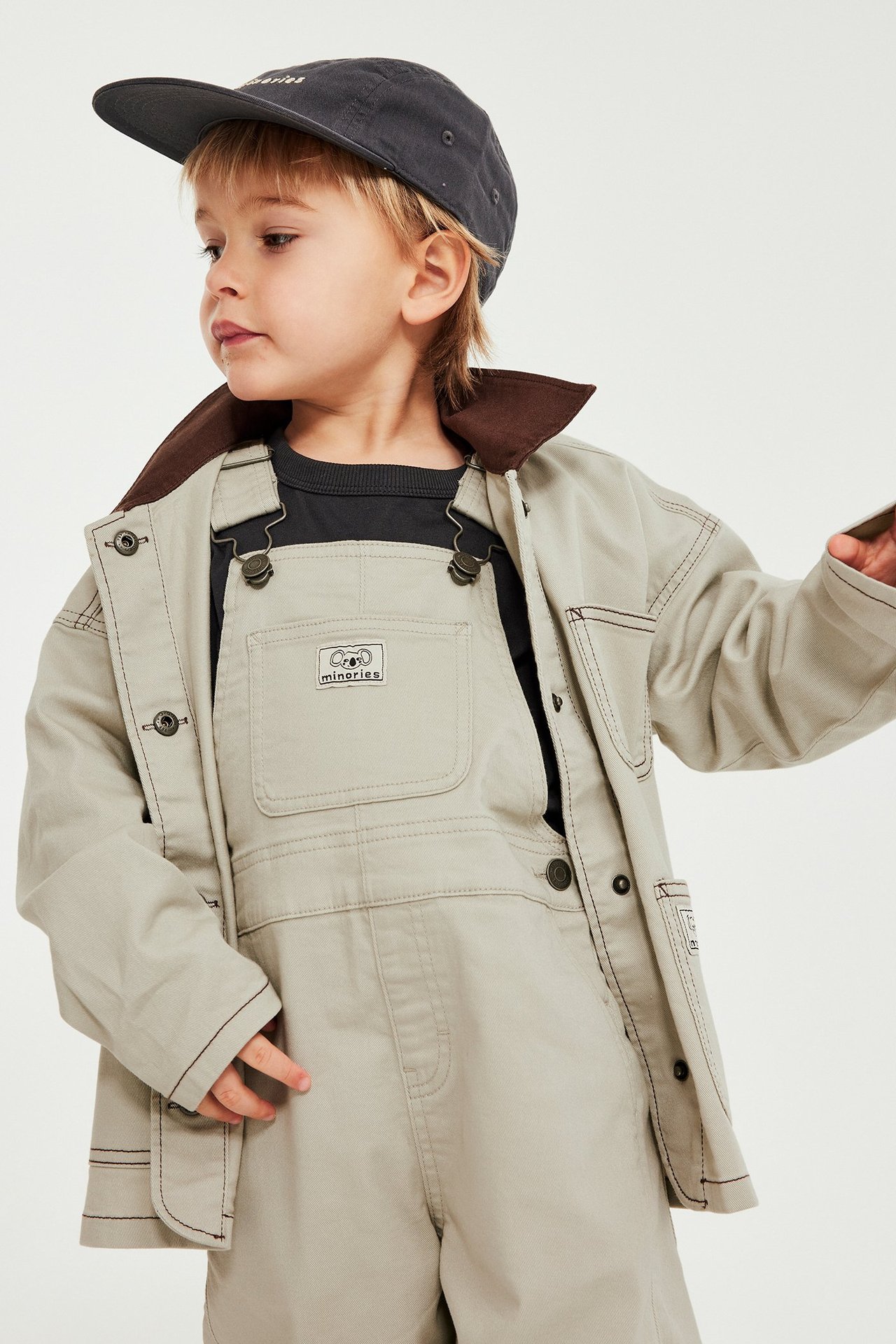 Lapsi edestä: beiget twill-henkselihousut mustan t-paidan päällä, beigen takin ja tummanharmaan lippalakin kanssa.