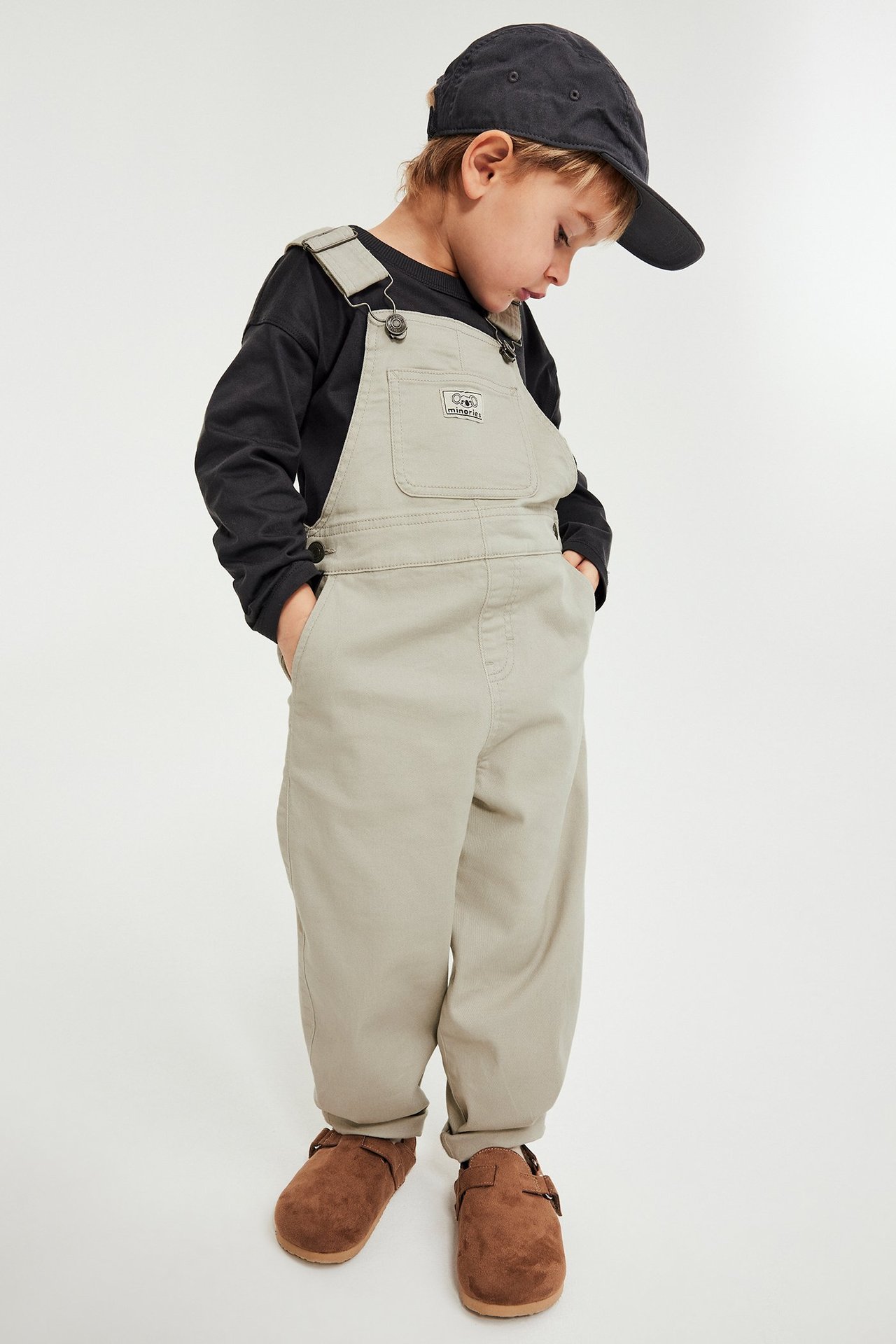 Lapsi beigenvärisissä twill-haalareissa, mustassa paidassa, mustassa lippiksessä, ruskeissa tohveleissa. Edestä.