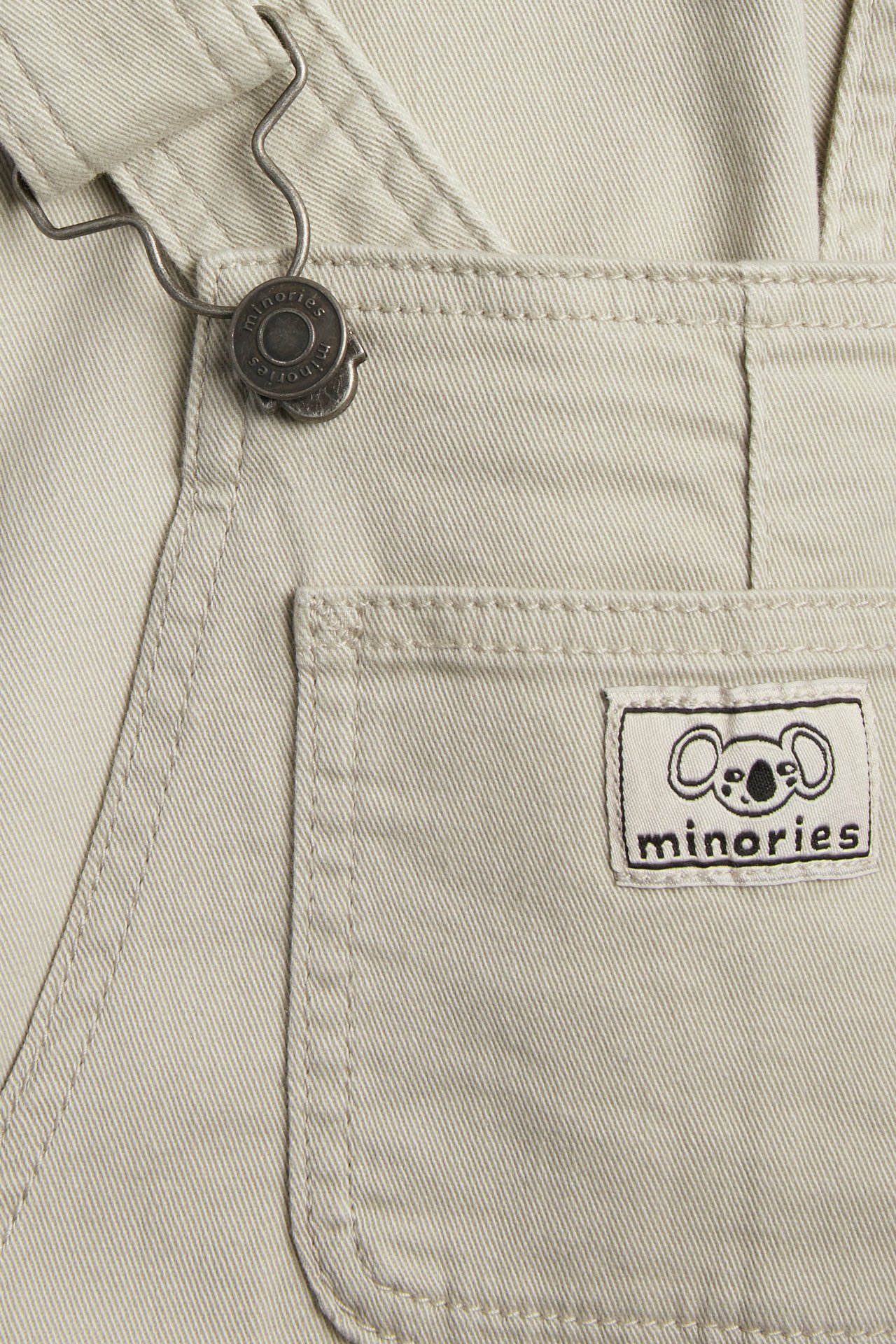 Lähikuva beigestä lasten twill-haalareista, joissa säädettävä olkain ja Minories-logomerkki.