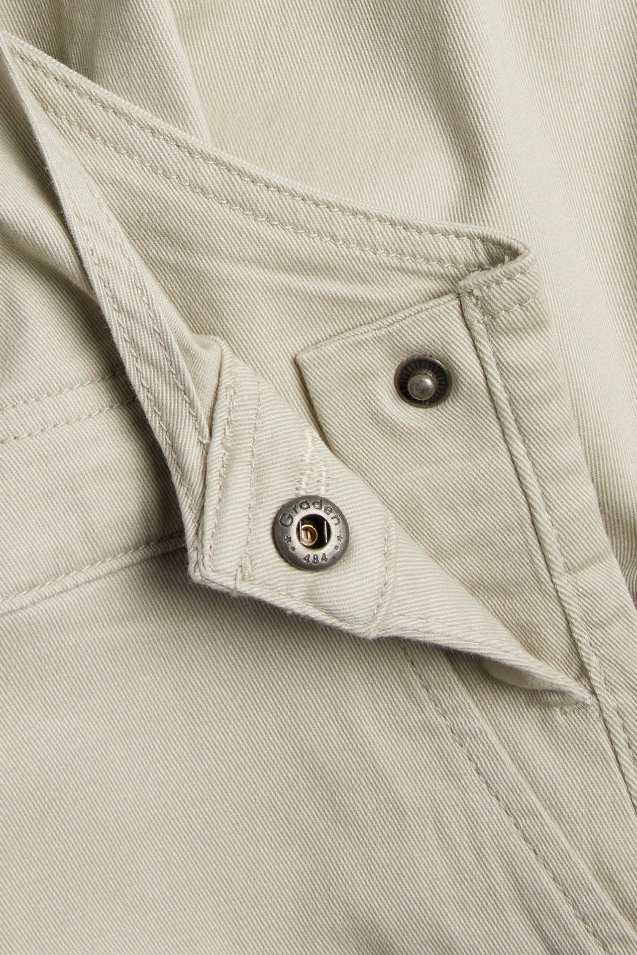 Lähikuva beigenvärisistä twill-haalarihousuista, näyttää metallipainonapin säädettävässä olkaimessa.