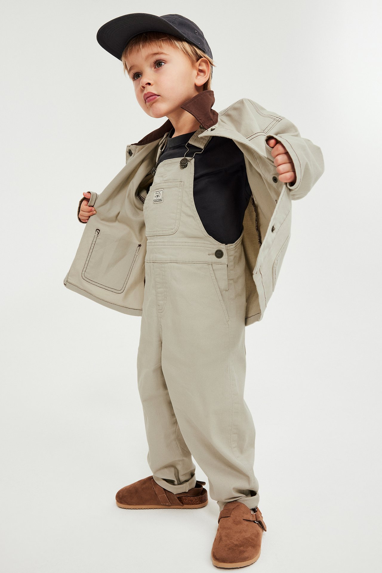 Edestä: Lapsi beigessä haalarissa, musta t-paita, avoin beige takki, harmaa lippis, ruskeat kengät.