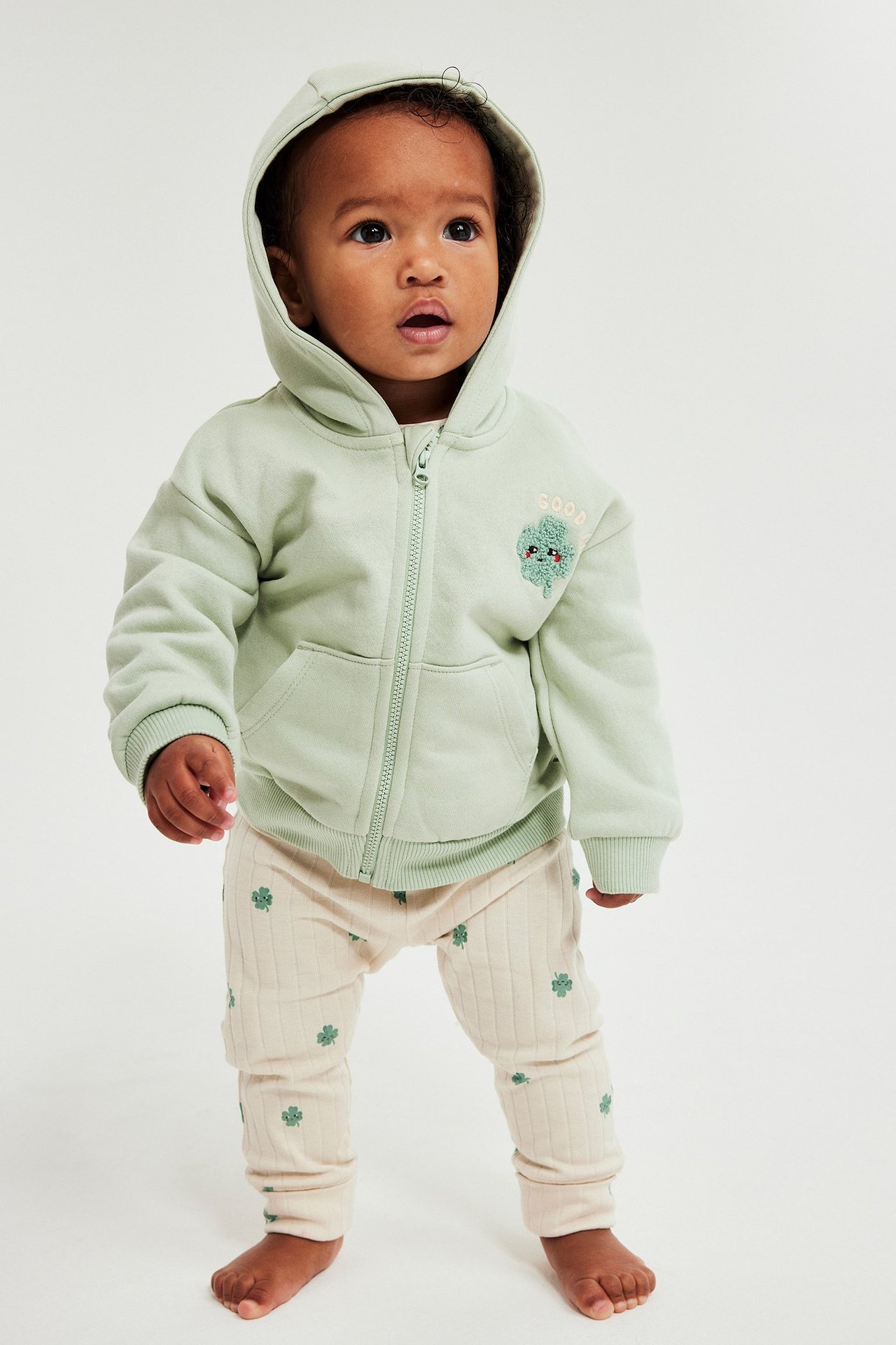 Edestä, vauva beigen ribatuissa apilakuvioisissa leggingseissä ja vaaleanvihreässä hupparissa.