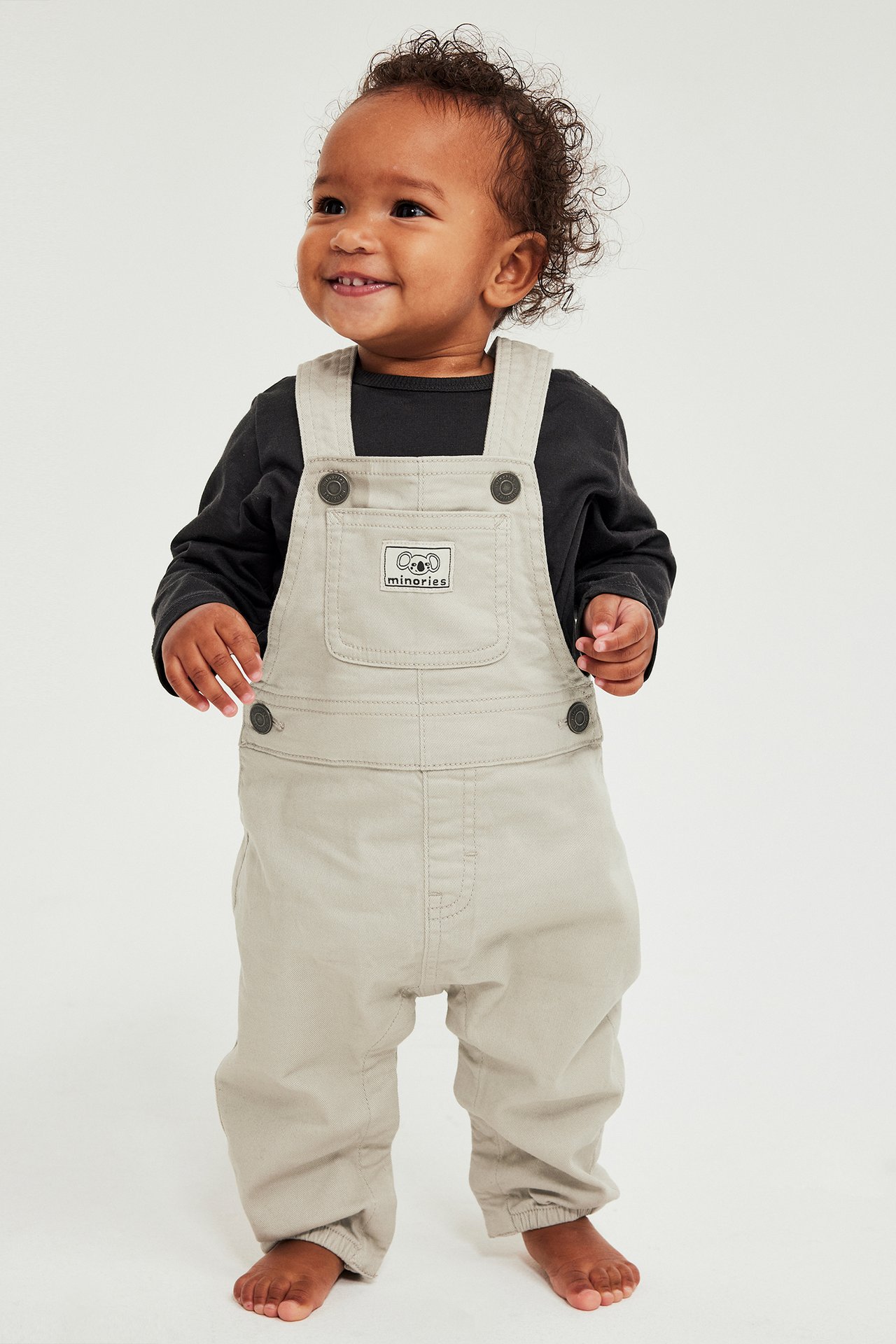 Beige twill-hängselhousut säädettävillä olkaimet ja Minories-logo edessä, lapsen malli.
