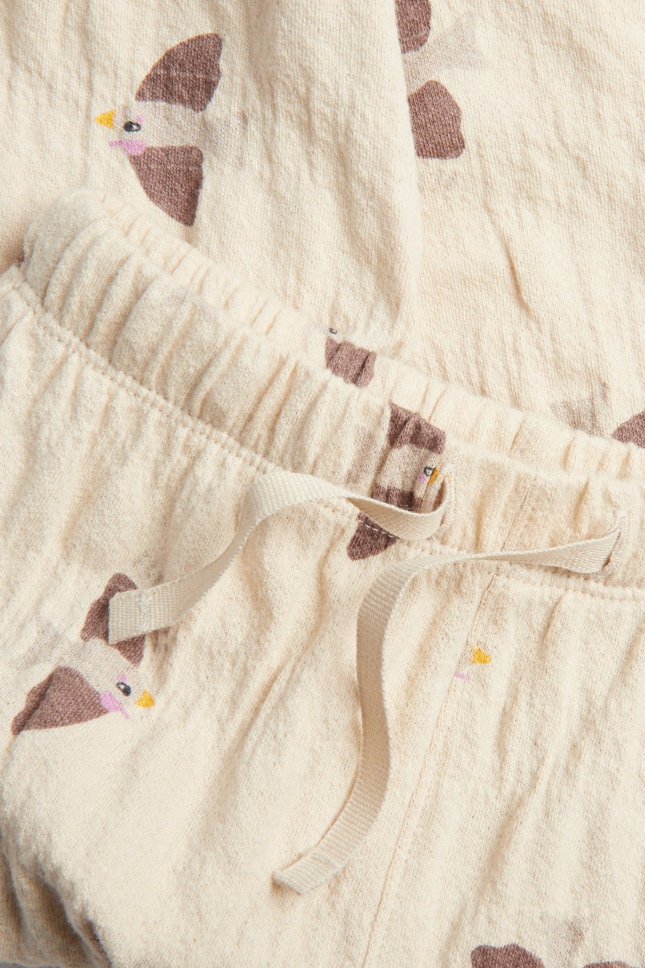 Nærbilde forfra av beige babyjoggebukser med fuglemønster, elastisk midje og snøring.