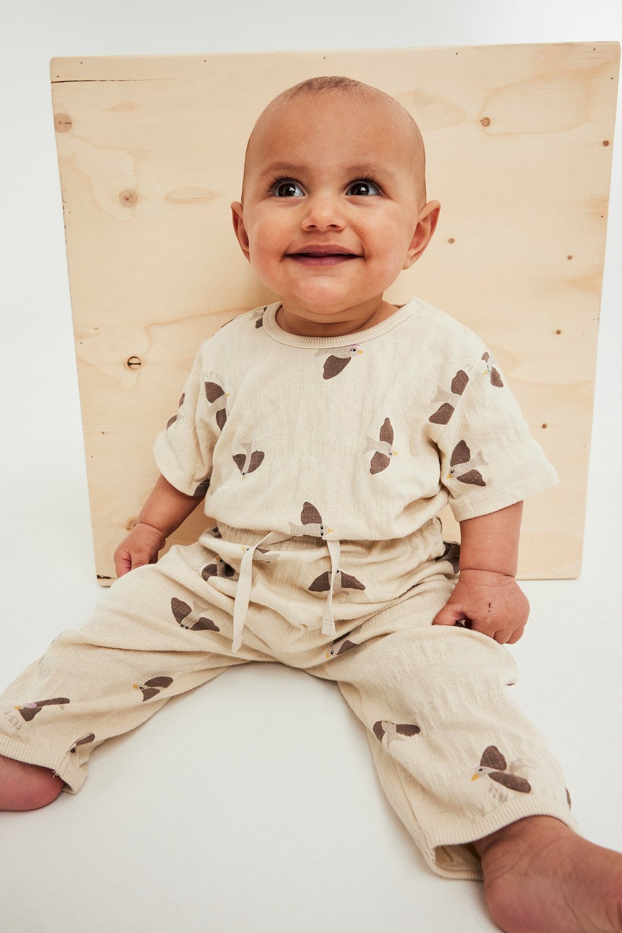 Baby i beige bomulls-joggesett med fuglemønster. Bukser med snøring og matchende kortermet topp.