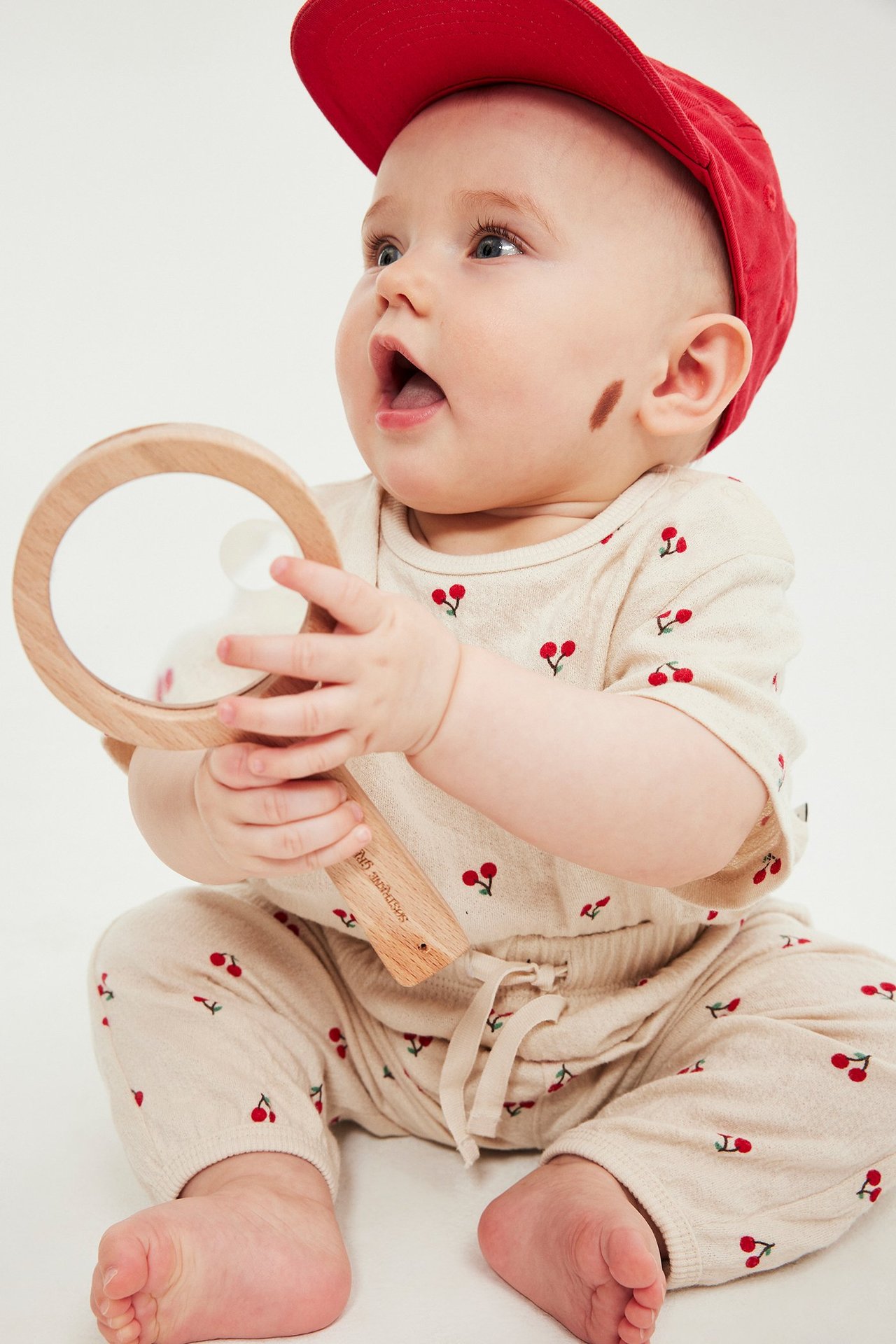 Widok z przodu: niemowlę w jasnobeżowych dresowych spodniach w wiśnie, z topem i czerwoną czapką, trzymające zabawkę.