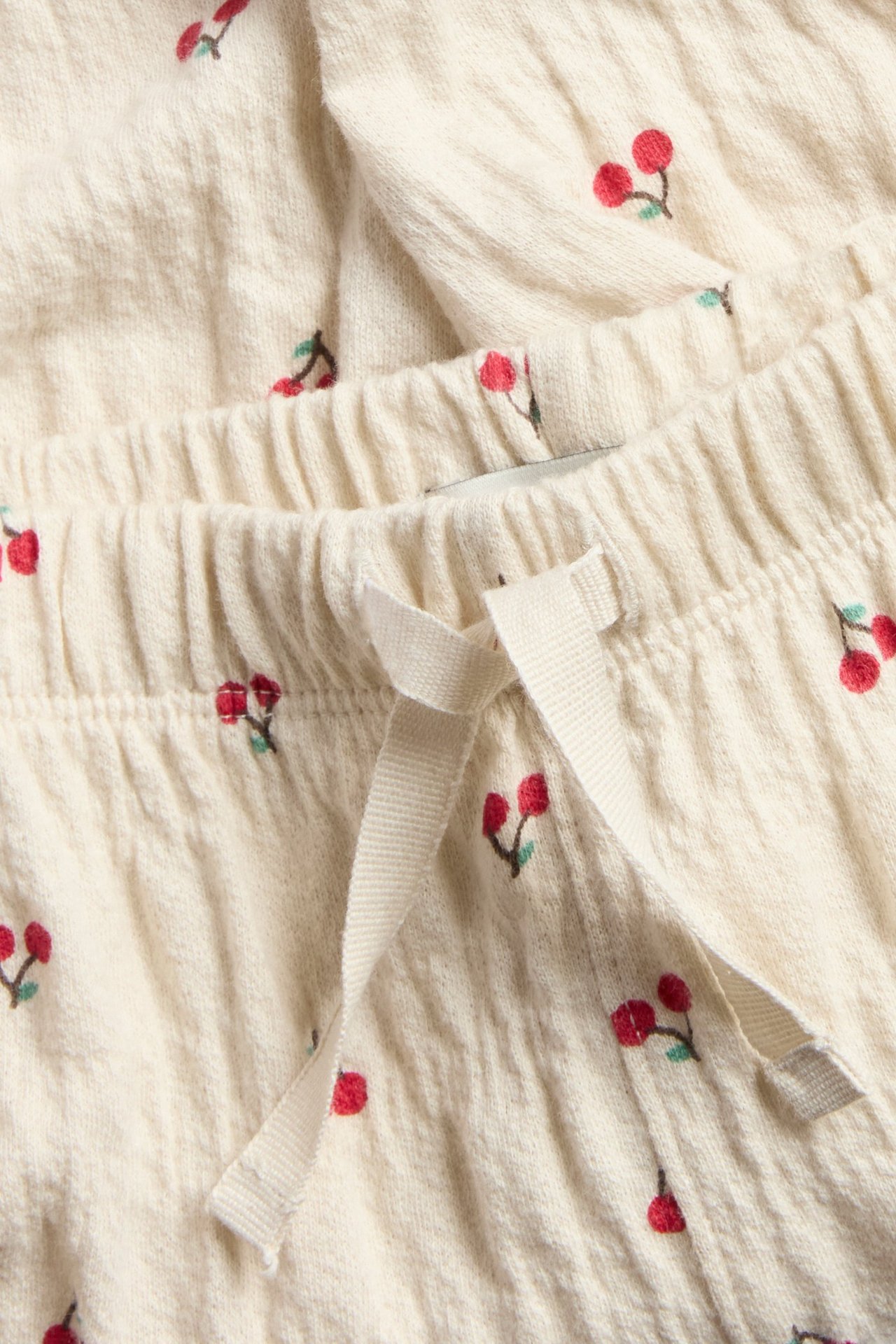 Närbild av ljusbeige babyjoggingbyxor med rött körsbärsmönster, resårmidja och dragsko.