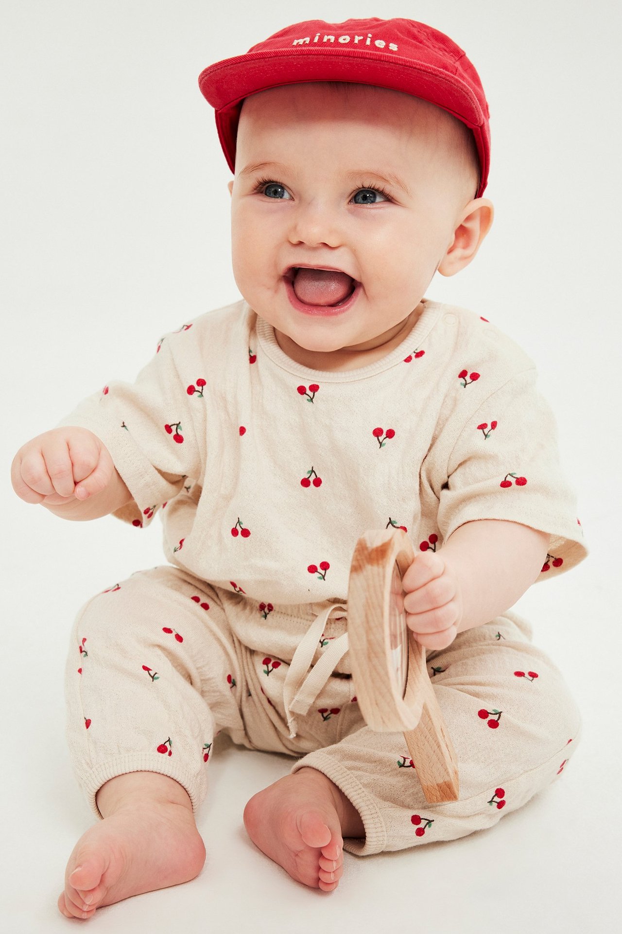 Uśmiechnięte niemowlę w beżowym dresie w wiśnie, czerwonej czapce, trzymające drewnianą zabawkę, widok z przodu.