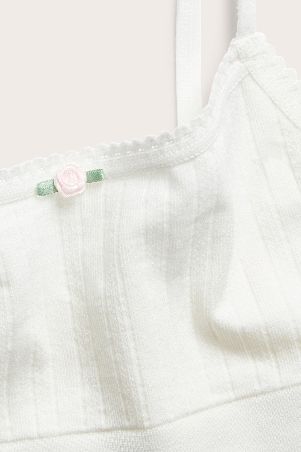 Zbliżenie na ecru top z dzianiny pointelle, z cienkimi ramiączkami, falbanką i różową różą.