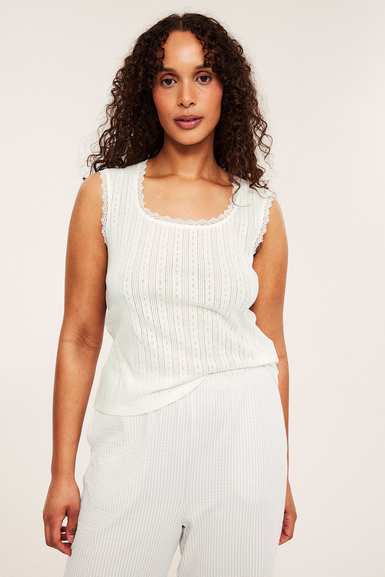 Frontbilde: kvinne i offwhite pointelle pyjamas topp med blondekant og lyse stripete pyjamasbukser.